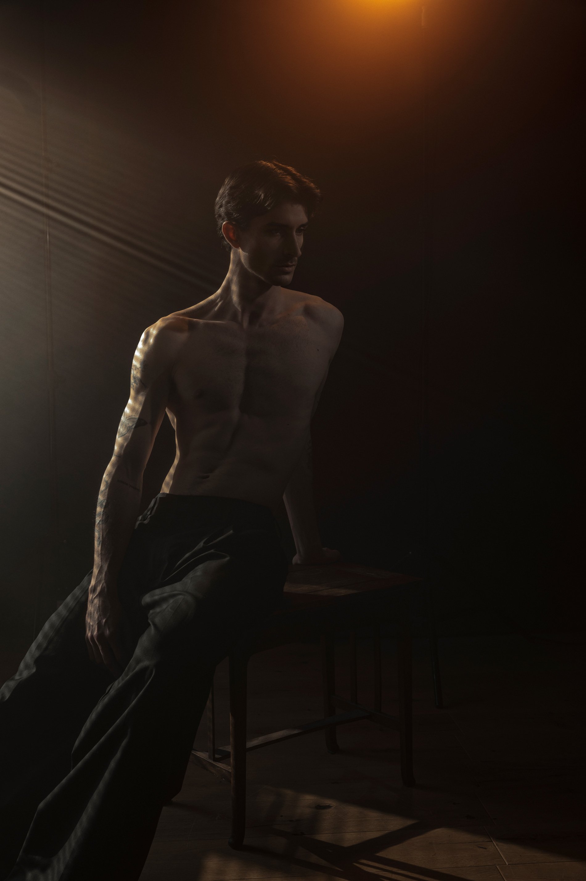 Ein junger, schmaler Mann sitzt nackt auf einem Stuhl in einem dunklen Raum, beleuchtet von warmem, seitlichem Licht.