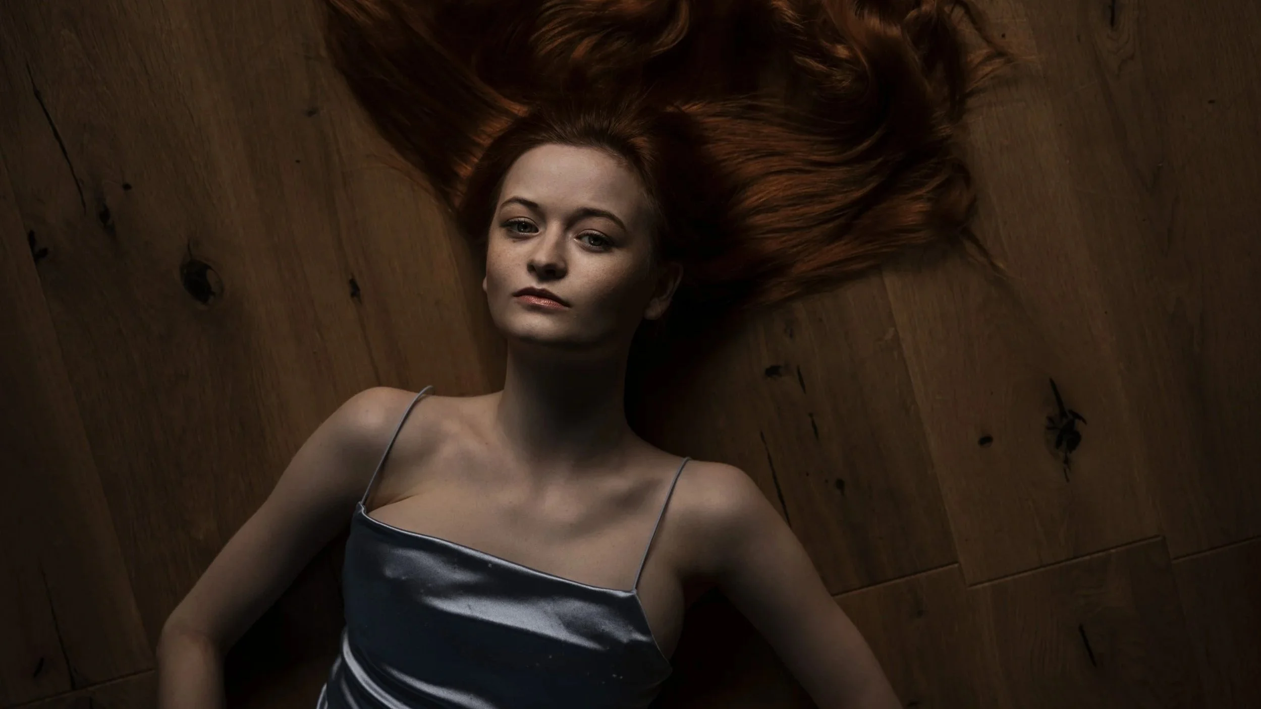 Junge Frau mit roten Haaren liegt auf einem Holzboden, schauen in die Kamera, trägt ein schwarzes Satin-Nachthemd.