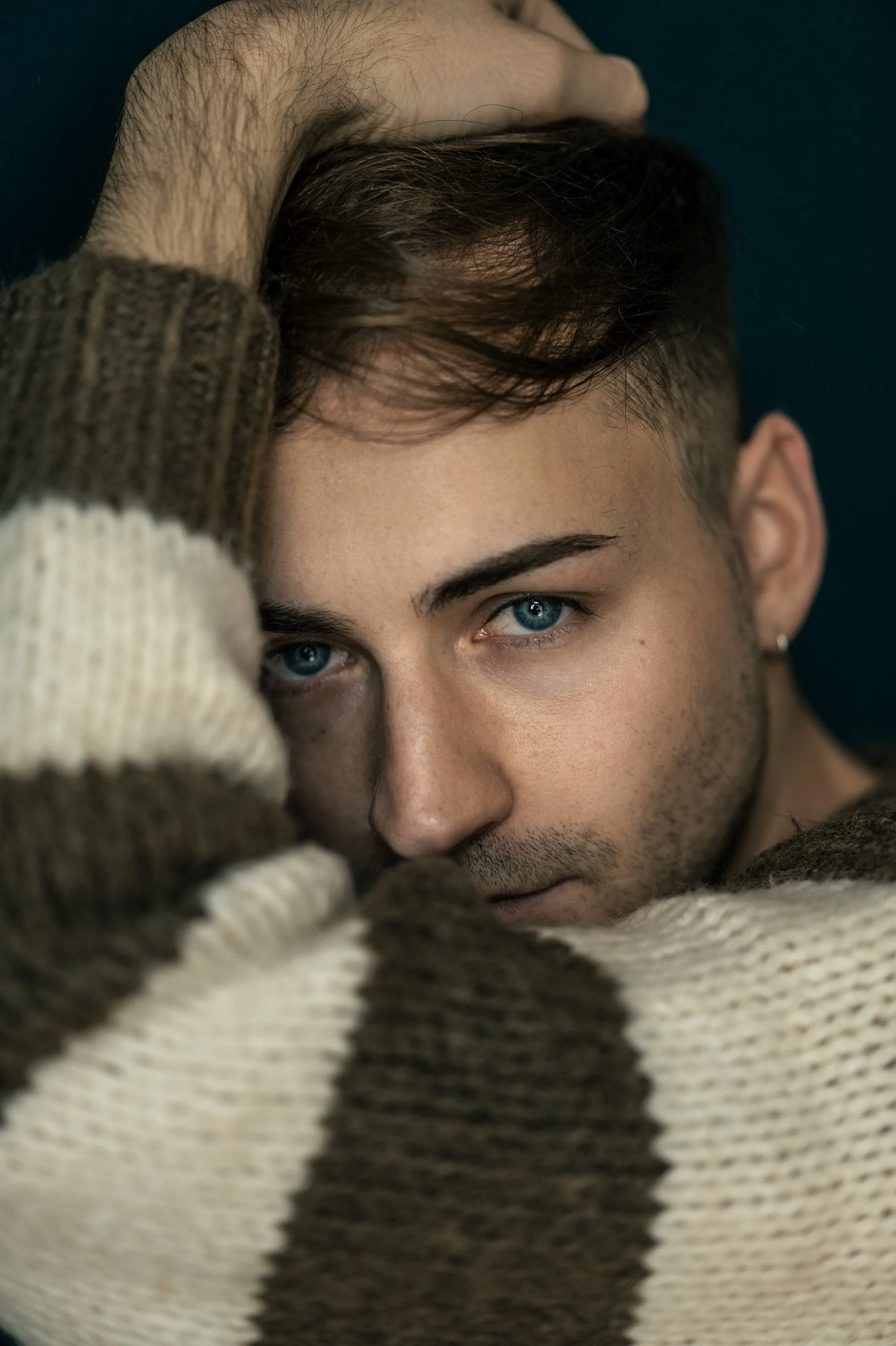 Nahaufnahme eines jungen Mannes mit blauen Augen, der eine braune und weiße gestrickte Jacke trägt und den Kopf leicht zur Seite neigt, während er in die Kamera schaut.