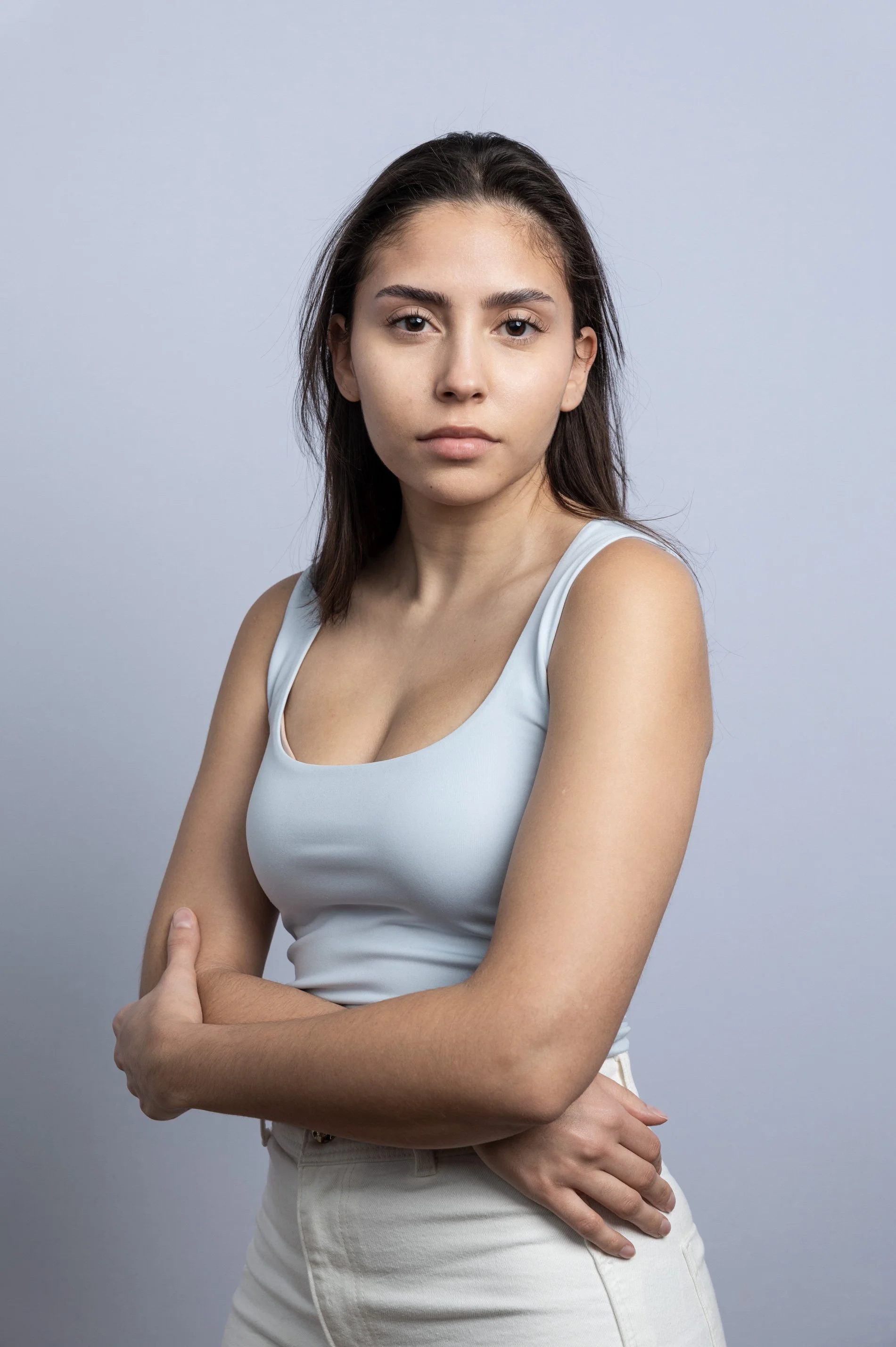 Junge Frau in weißem Tanktop und weißen Hosen steht vor neutralem grauem Hintergrund, Blick gerade, Arme verschränkt.
