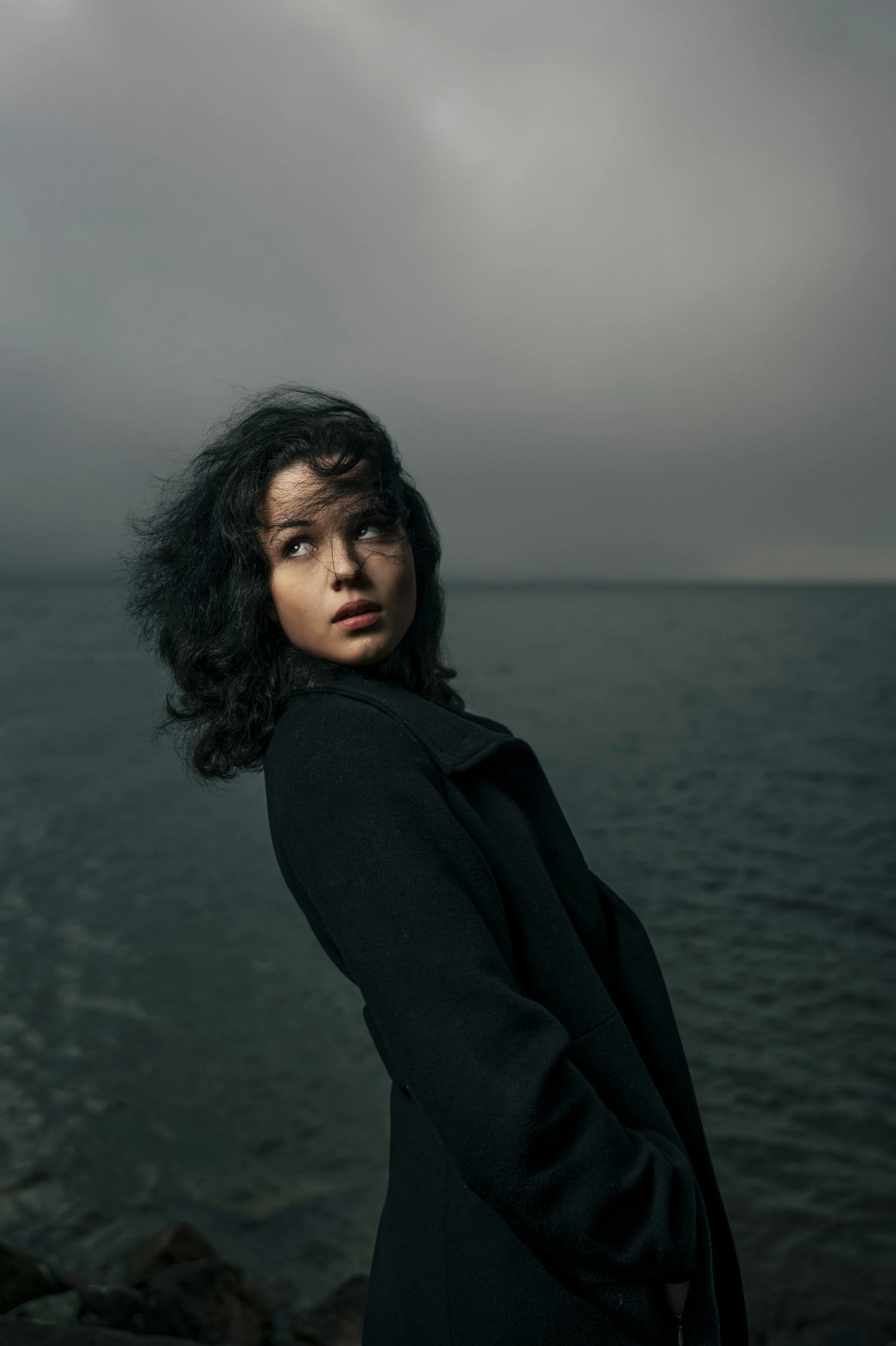 Junge Frau mit schwarzen, losen Haaren steht am Wasser bei bewölktem Himmel, trägt schwarzen Mantel, blickt nach oben und zur Seite.