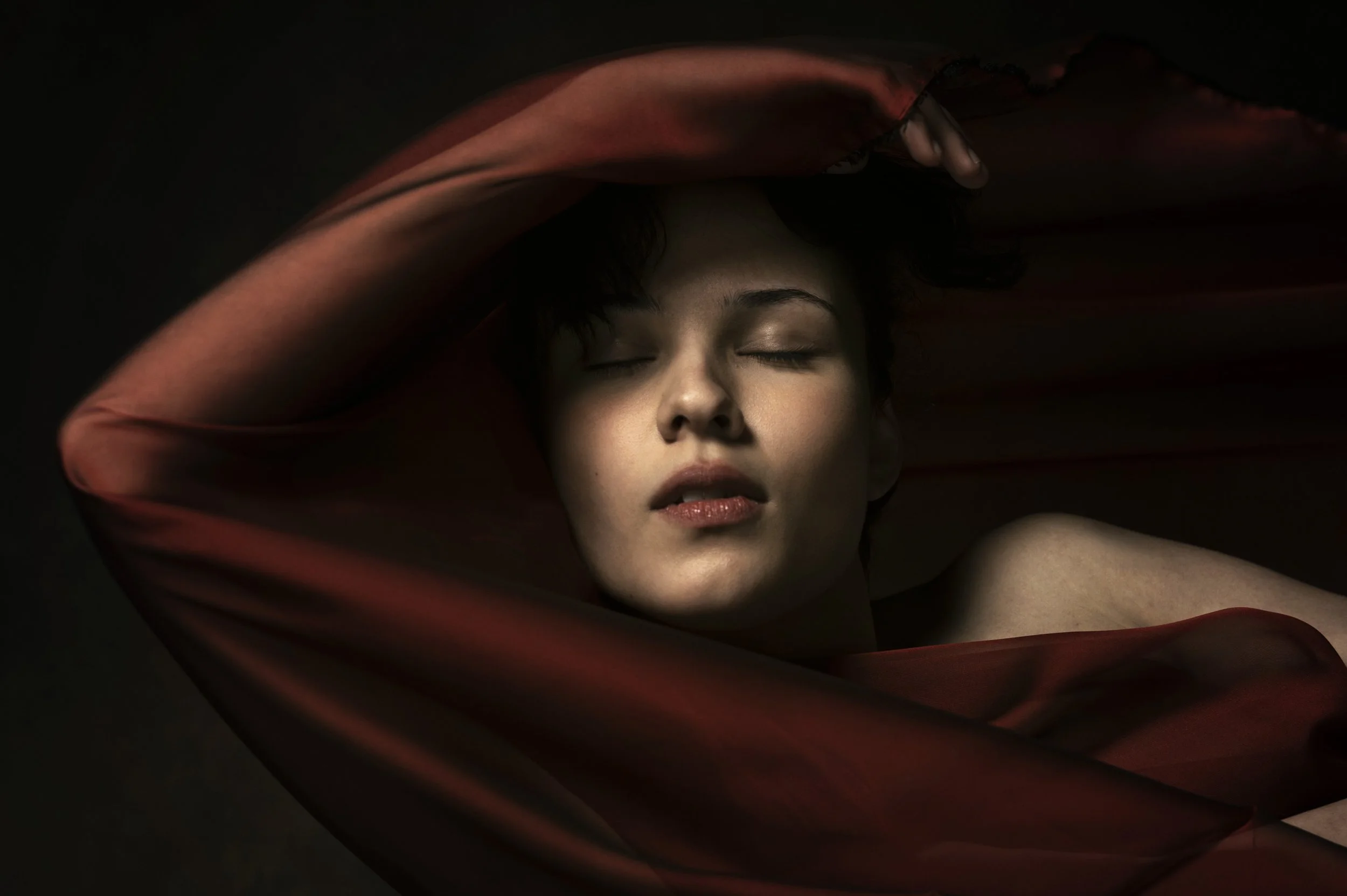 Nahaufnahme eines Gesichts einer Frau mit geschlossenen Augen, umrahmt von einem roten, transparenten Stoff, der ihre Arme und Schultern umhüllt, vor dunklem Hintergrund.