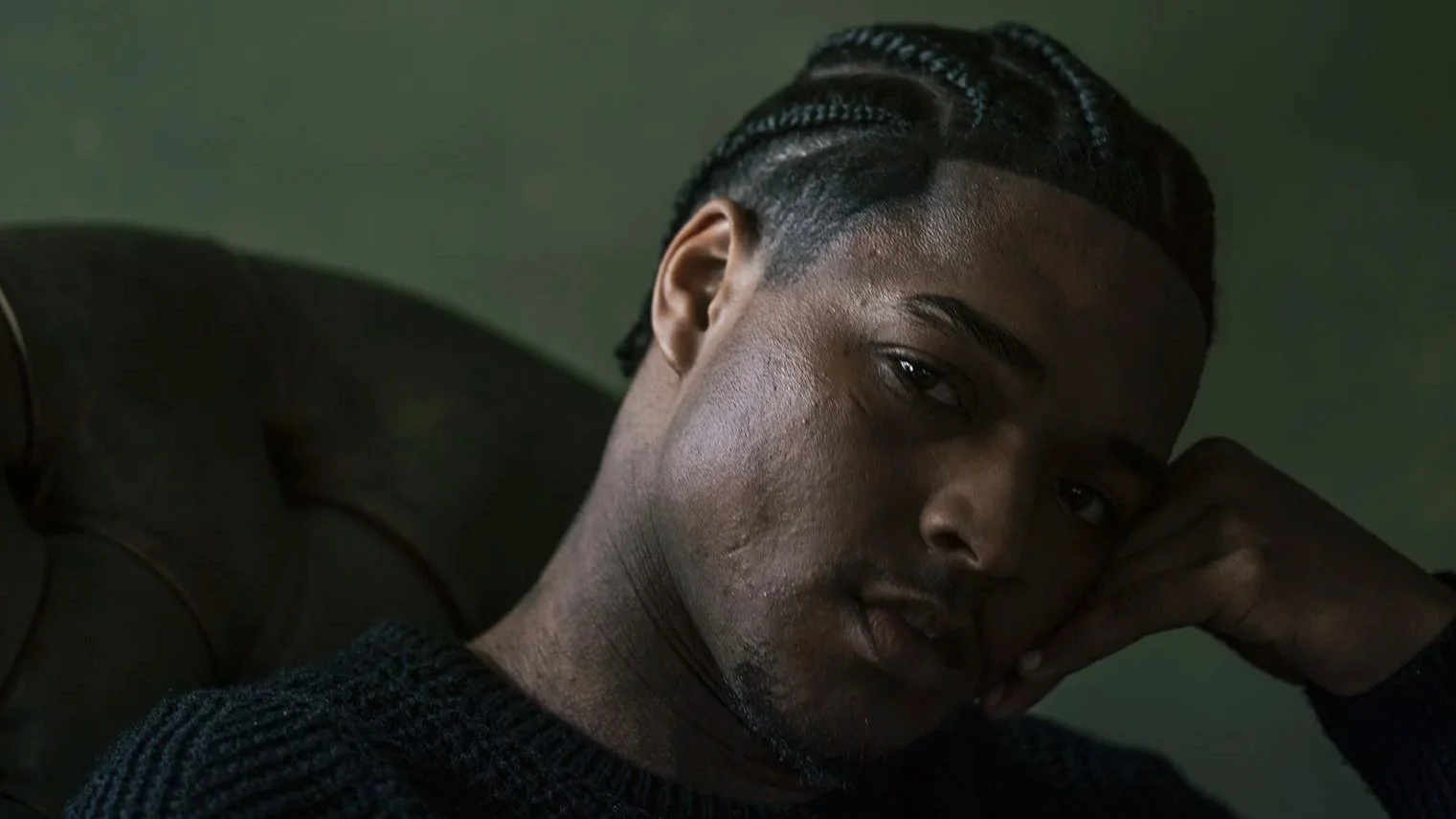 Nahaufnahme eines Mannes mit sprudelnden, cornrow-ähnlichen Frisuren, die an den Seiten kurz geschnitten sind, liegt auf einem grauen Kissen, trägt einen schwarzen Pullover und blickt nach oben.