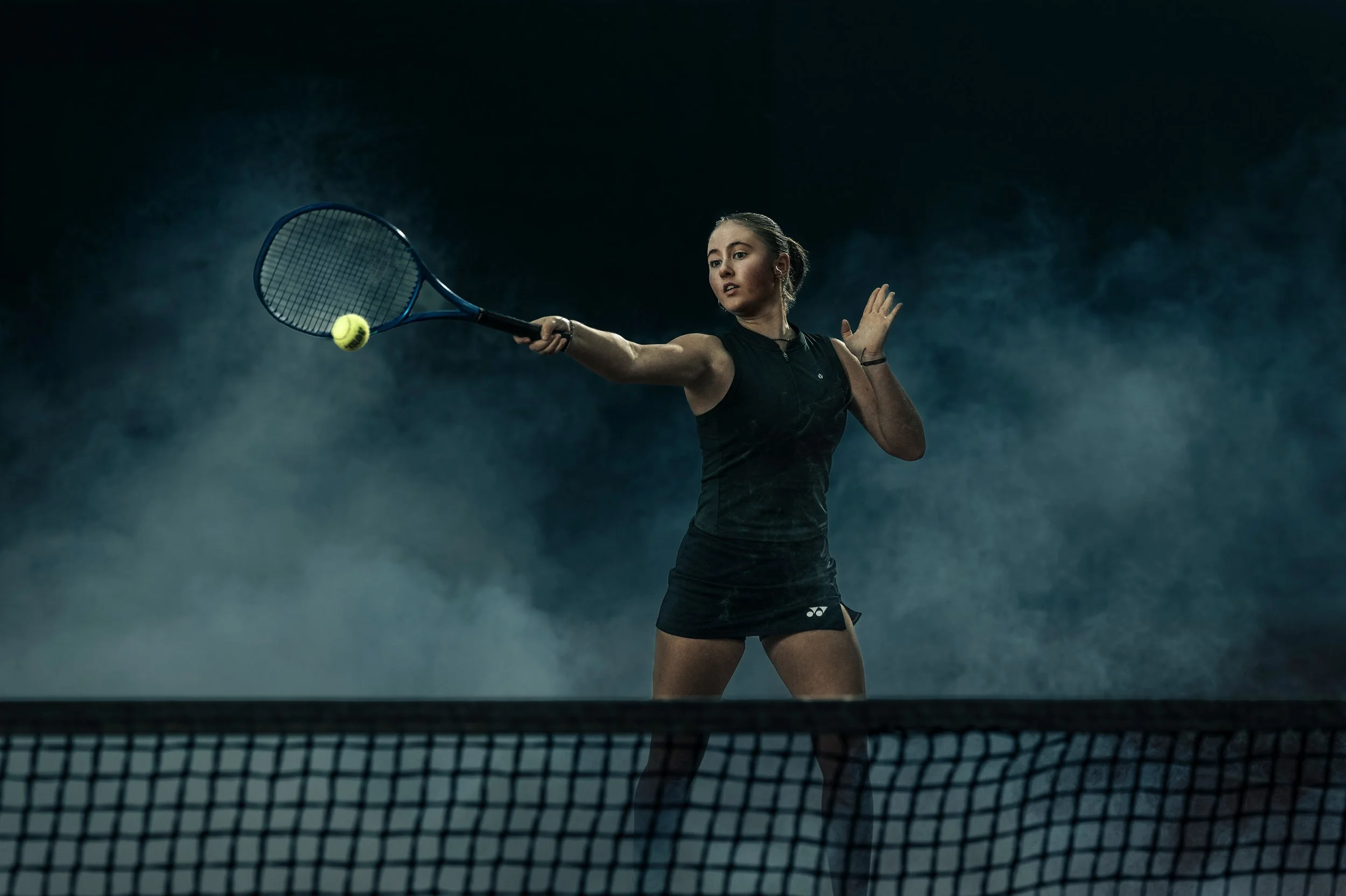 Tennisspielerin im schwarzen Sportoutfit beim Schlag auf einem Tennisspielplatz mit dunklem Hintergrund und Nebel.
