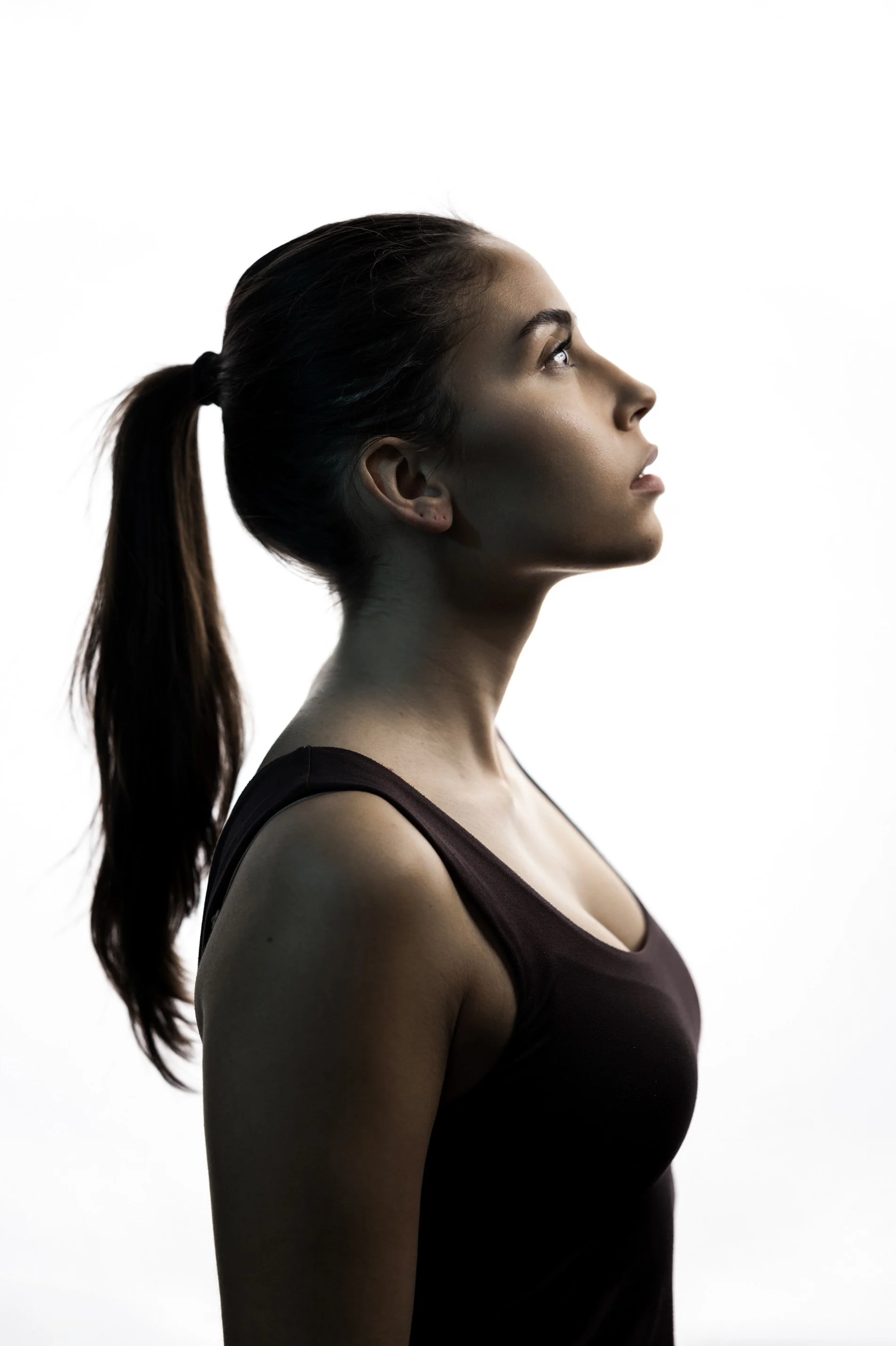 Seitenansicht einer jungen Frau mit dunklem Haar in Pferdeschwanz, die nach oben schaut, vor weißem Hintergrund.