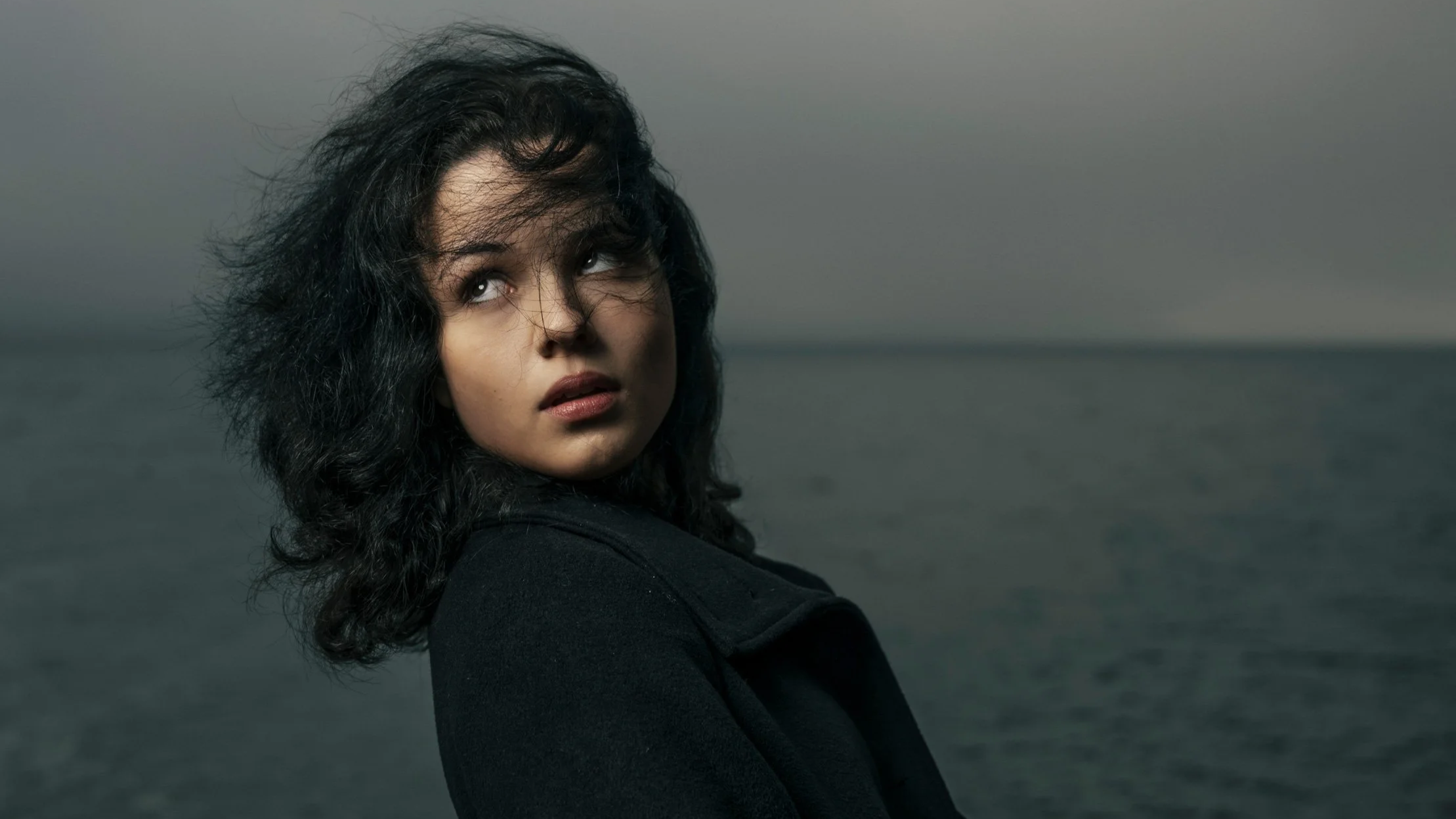 Junge Frau mit dunklen, lockigen Haaren schaut nach oben an einem Gewässer bei bewölktem Himmel.
