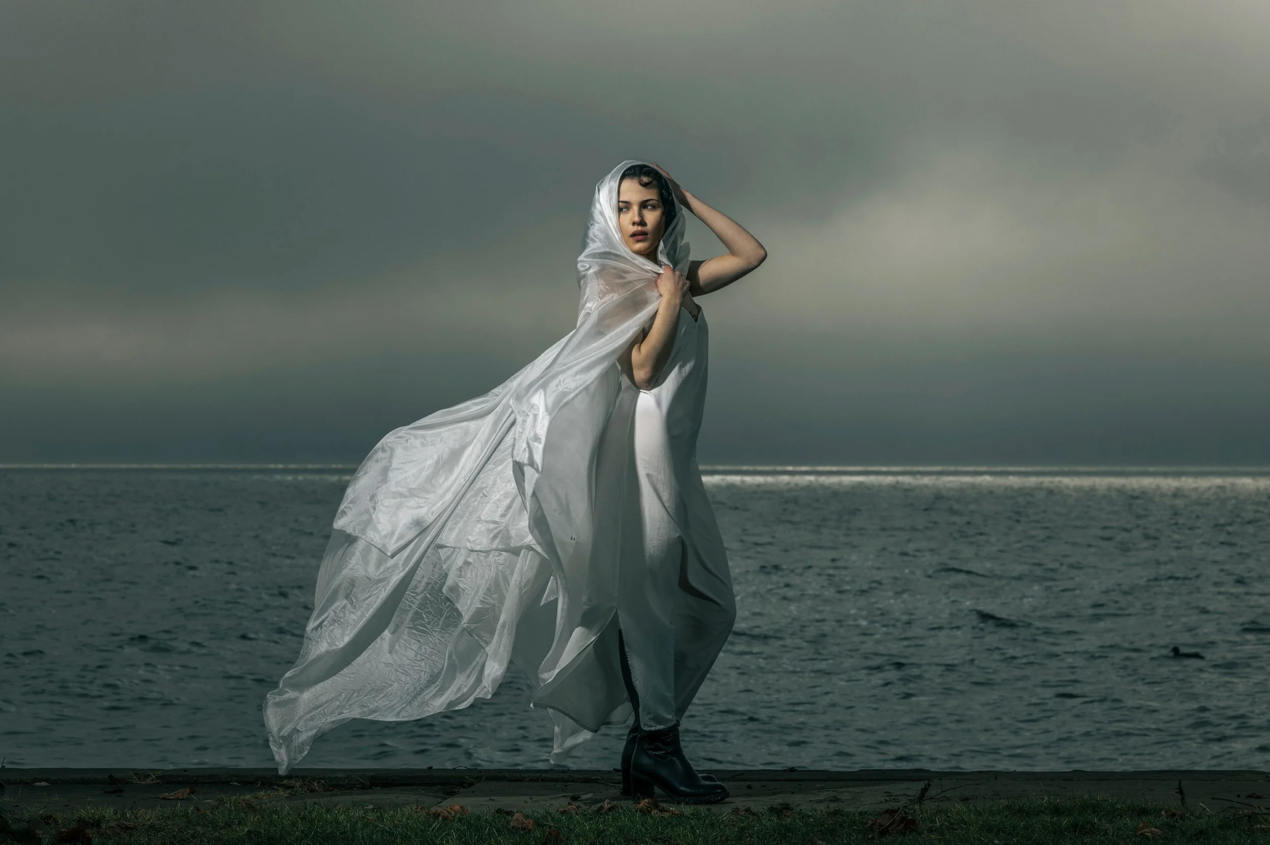 Frau steht am Meer, trägt ein weißes Kleid und einen silbernen Umhang, schaut nach links, bei bewölktem Himmel.