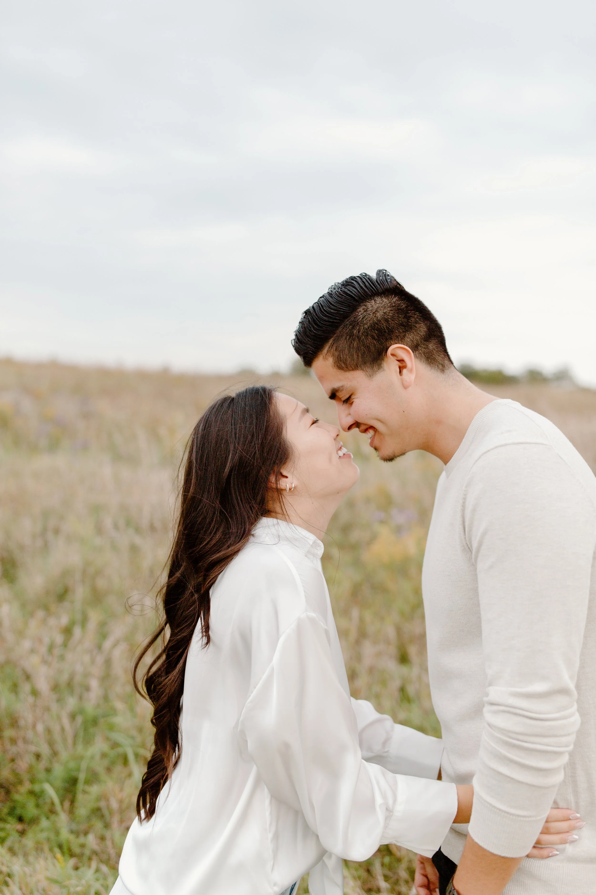 A couple standing close with their foreheads touching and smiling at each other outdoors in a grassy field.