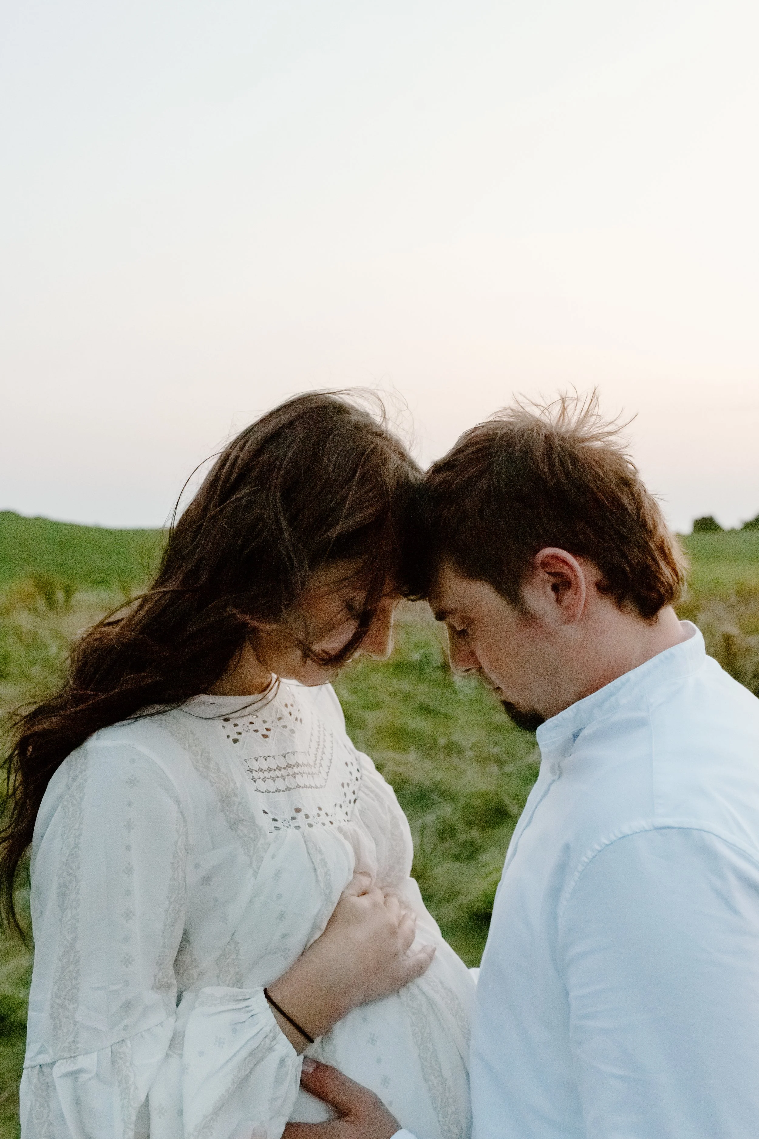 A couple with a pregnant woman and a man standing close with foreheads touching outdoors at sunset.