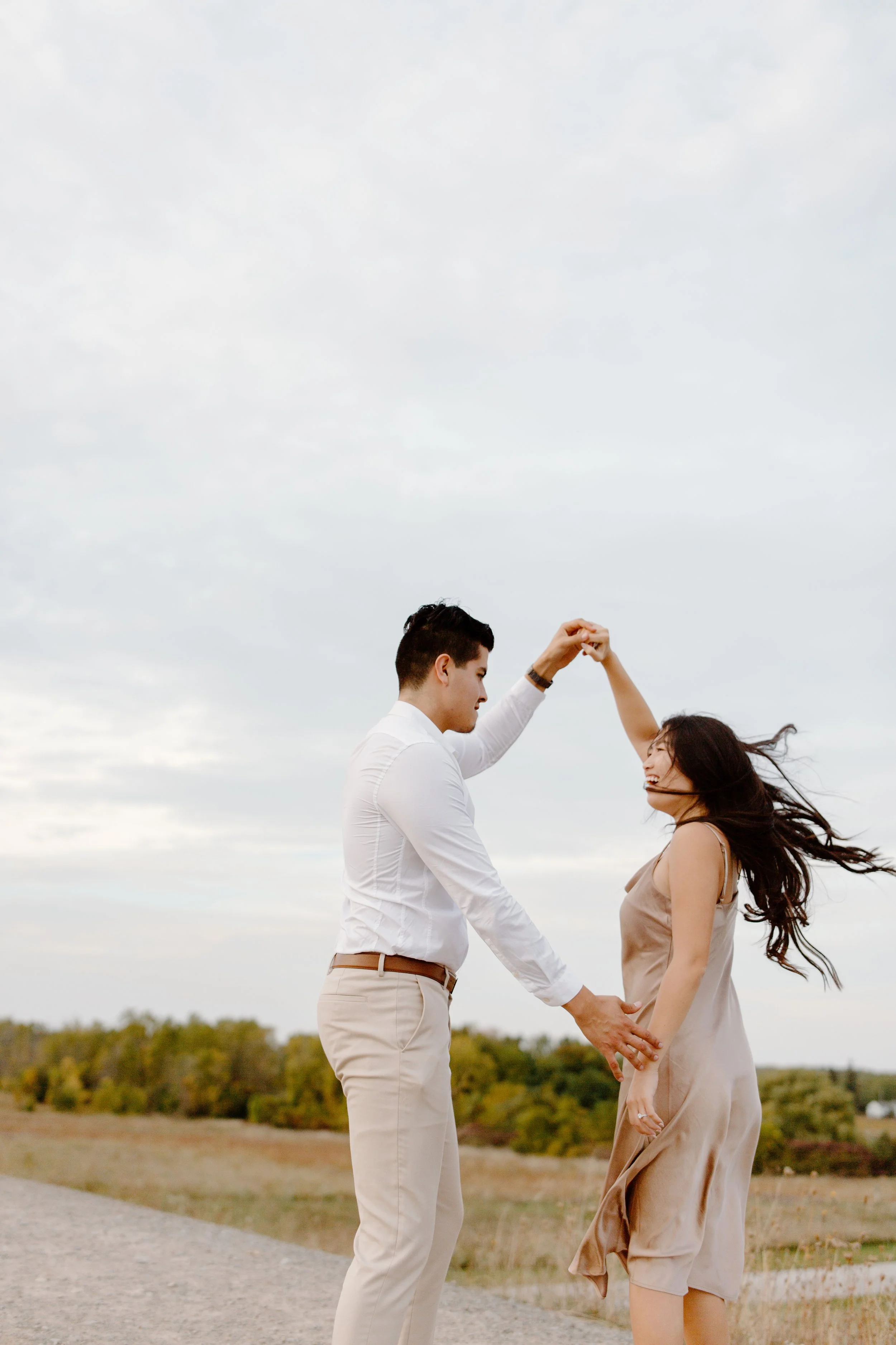 A man and woman dancing outdoors on a grassy field with trees. The man is in a white shirt and beige pants, and the woman is in a light beige dress. They are holding hands and twirling, smiling and enjoying each other's company.