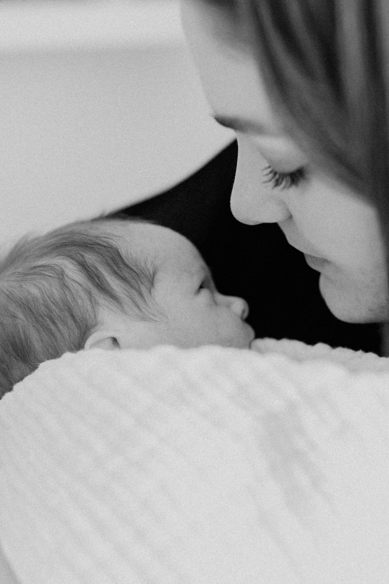 Close-up black and white photo of a woman and a baby face-to-face, gently touching noses, with the baby lying on a soft surface.