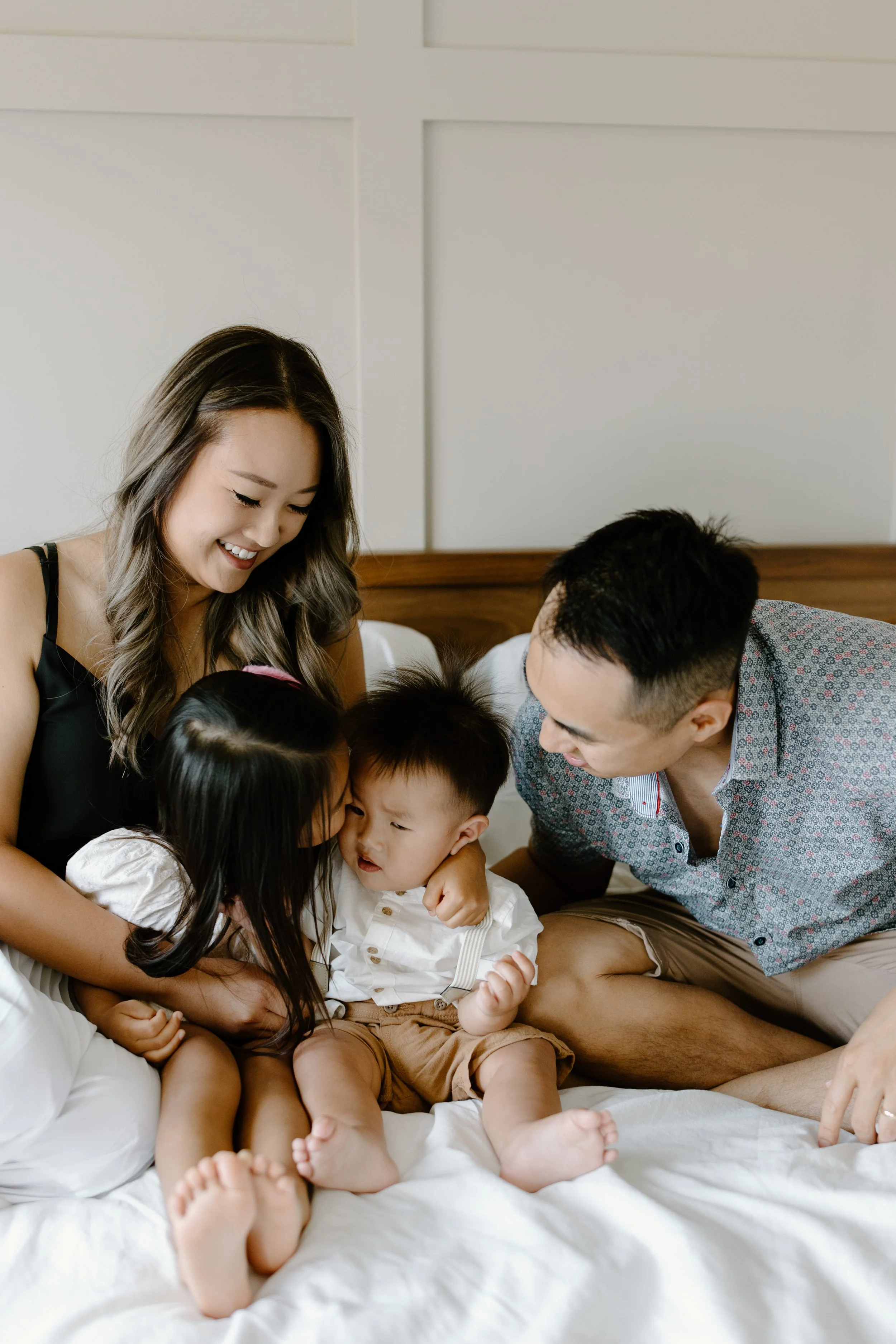 A happy Asian family of four sitting on a bed, smiling and playing together. The mother has long brown hair, and the father has short black hair. The two children, a girl and a boy, sit between their parents, with the girl giving the boy a kiss on th