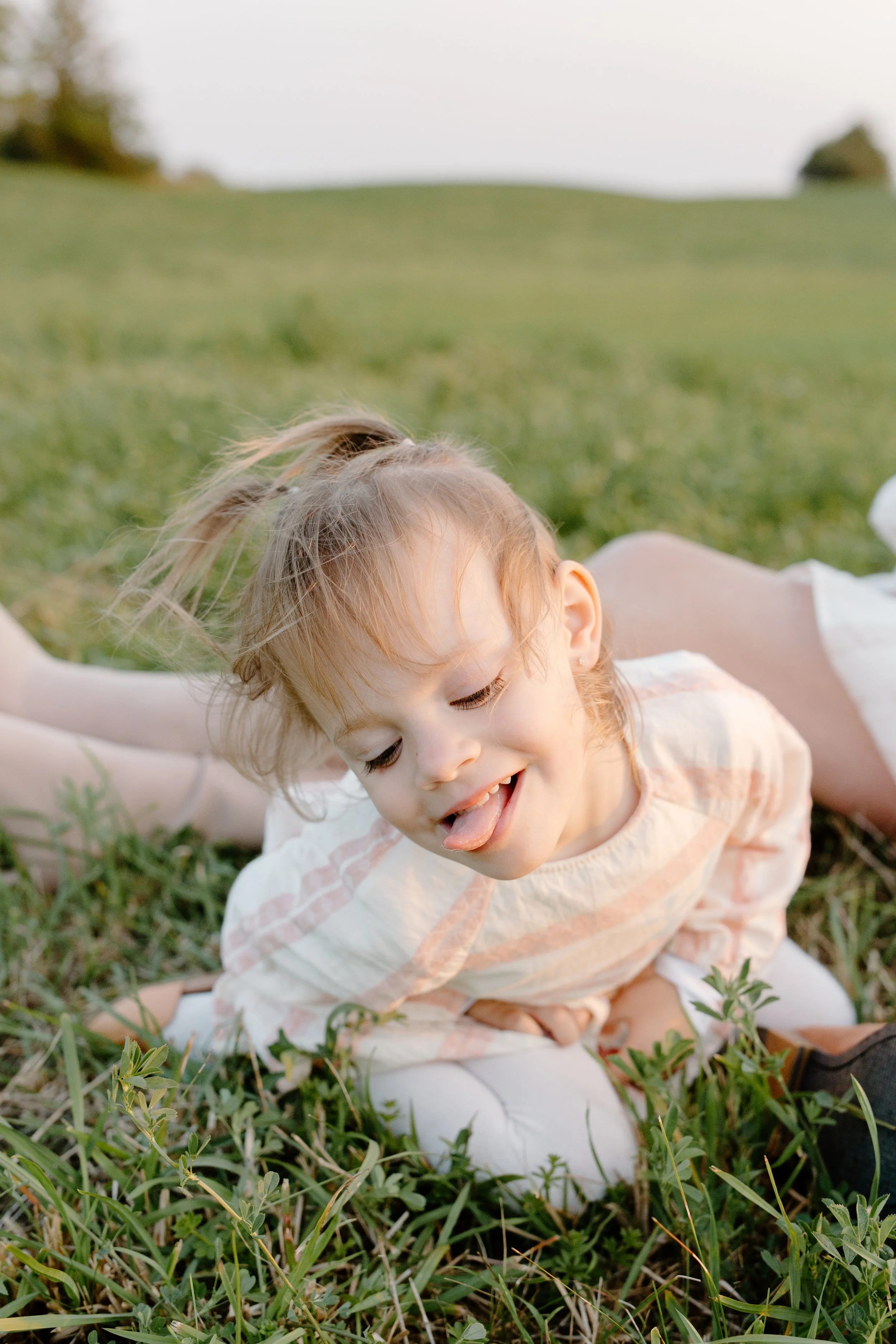 Young girl with light brown hair tied in a ponytail, laughing and sticking out her tongue, lying on green grass outdoors.