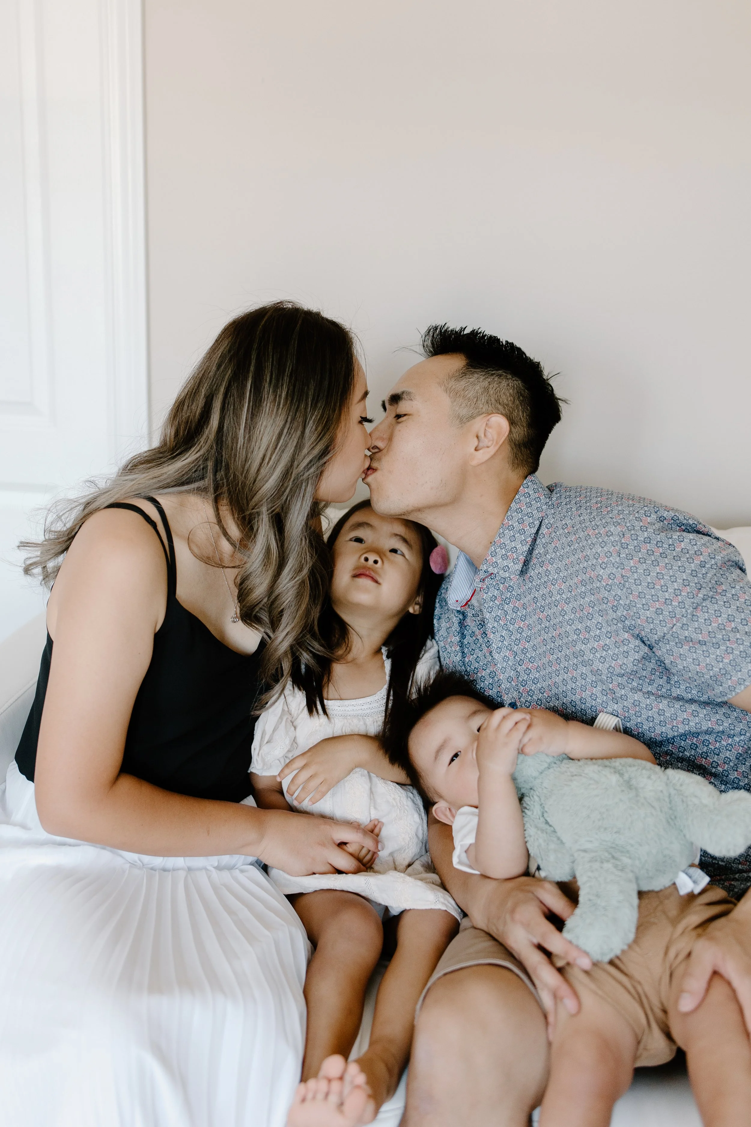 A family of four sitting on a couch, with two parents kissing each other and two children, a girl and a boy, looking at them, with the girl sitting between the parents and the boy lying on the father's lap holding a stuffed animal.