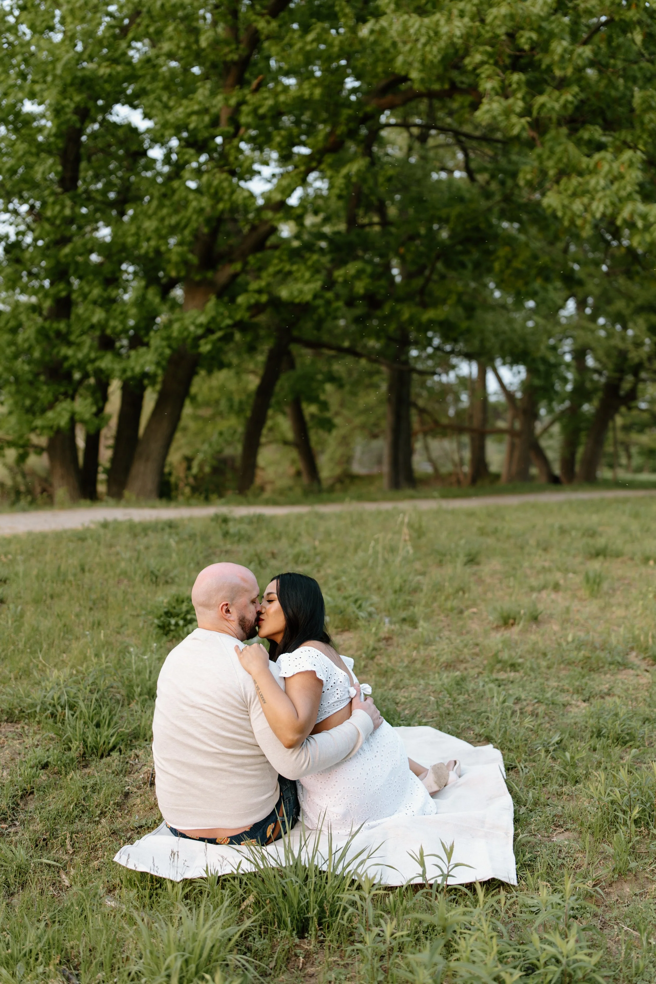 A couple kisses on a white blanket on grass in a park, with trees in the background.