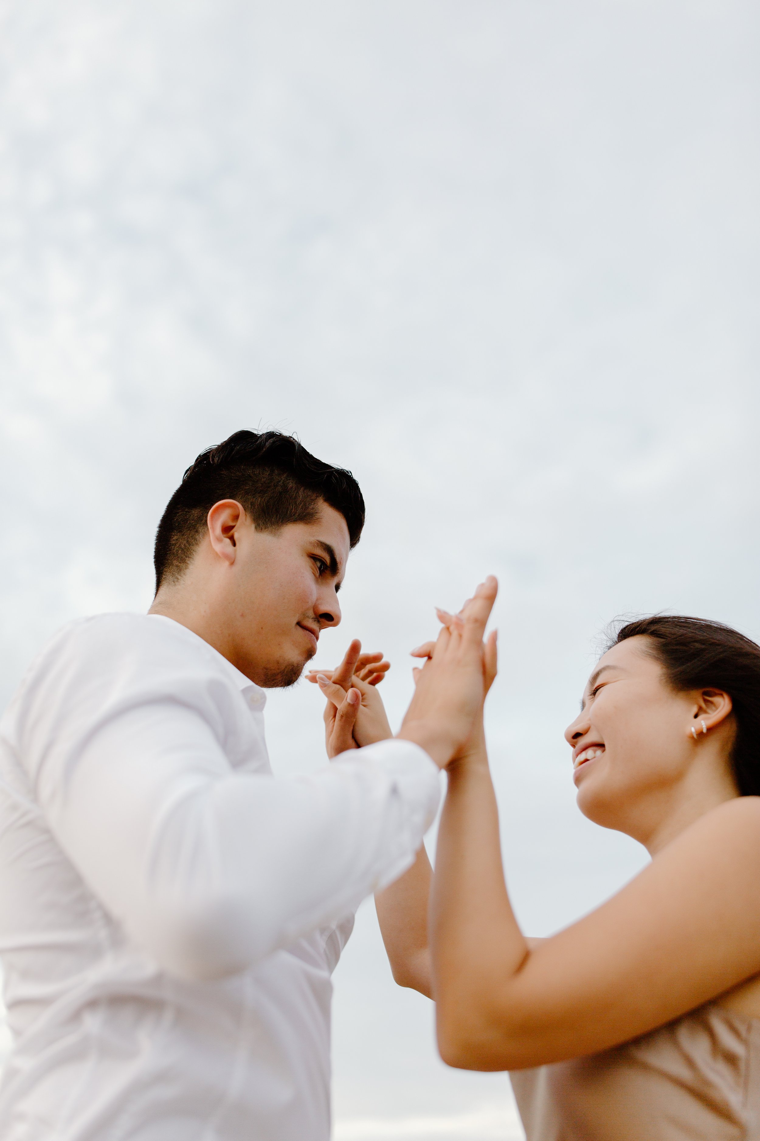 A man and woman smiling and touching hands during a playful moment outdoors against a cloudy sky.