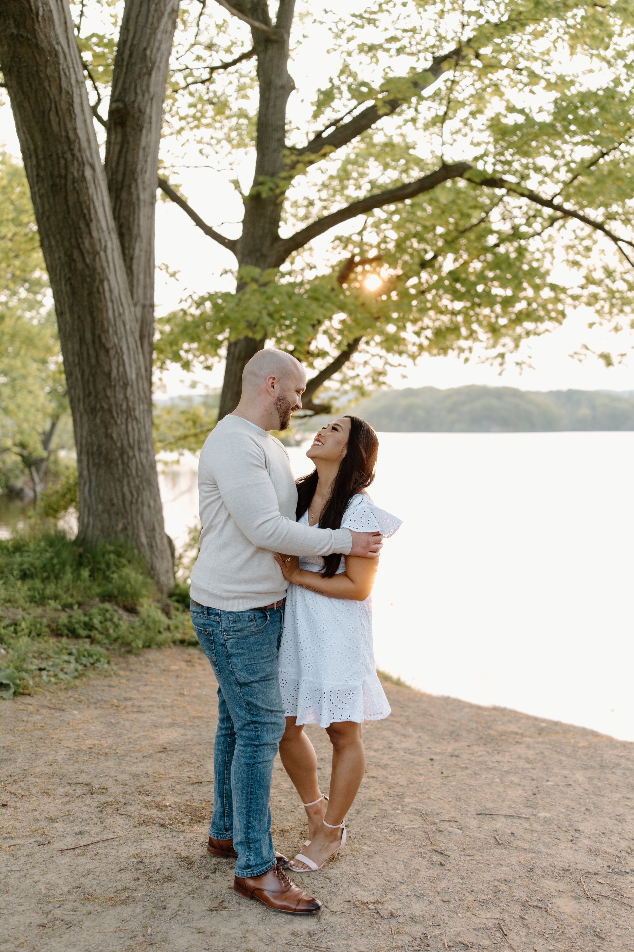 A couple embracing near a lake, standing under a large tree with sunlight filtering through the leaves during sunset.