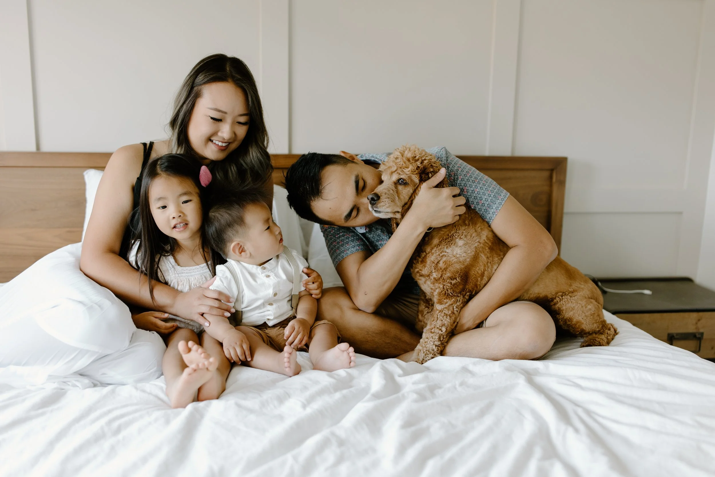 Family of four with two children and a man playing with a golden retriever puppy on a bed in a bedroom.