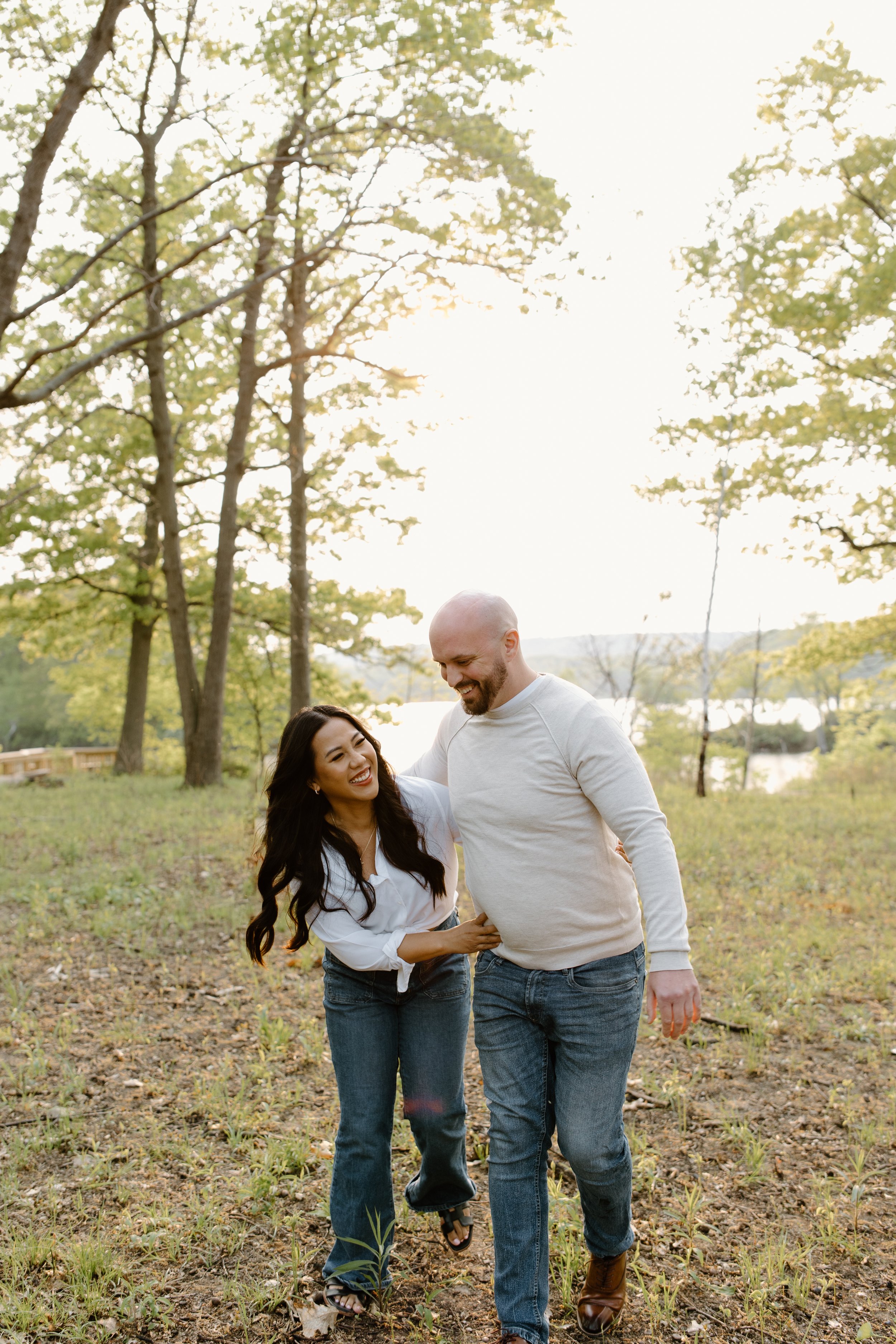 A couple walking in a park during the daytime, smiling and enjoying each other's company. The woman has long dark hair, and the man is bald with a beard. They are dressed casually in jeans and light tops, surrounded by trees and natural scenery.