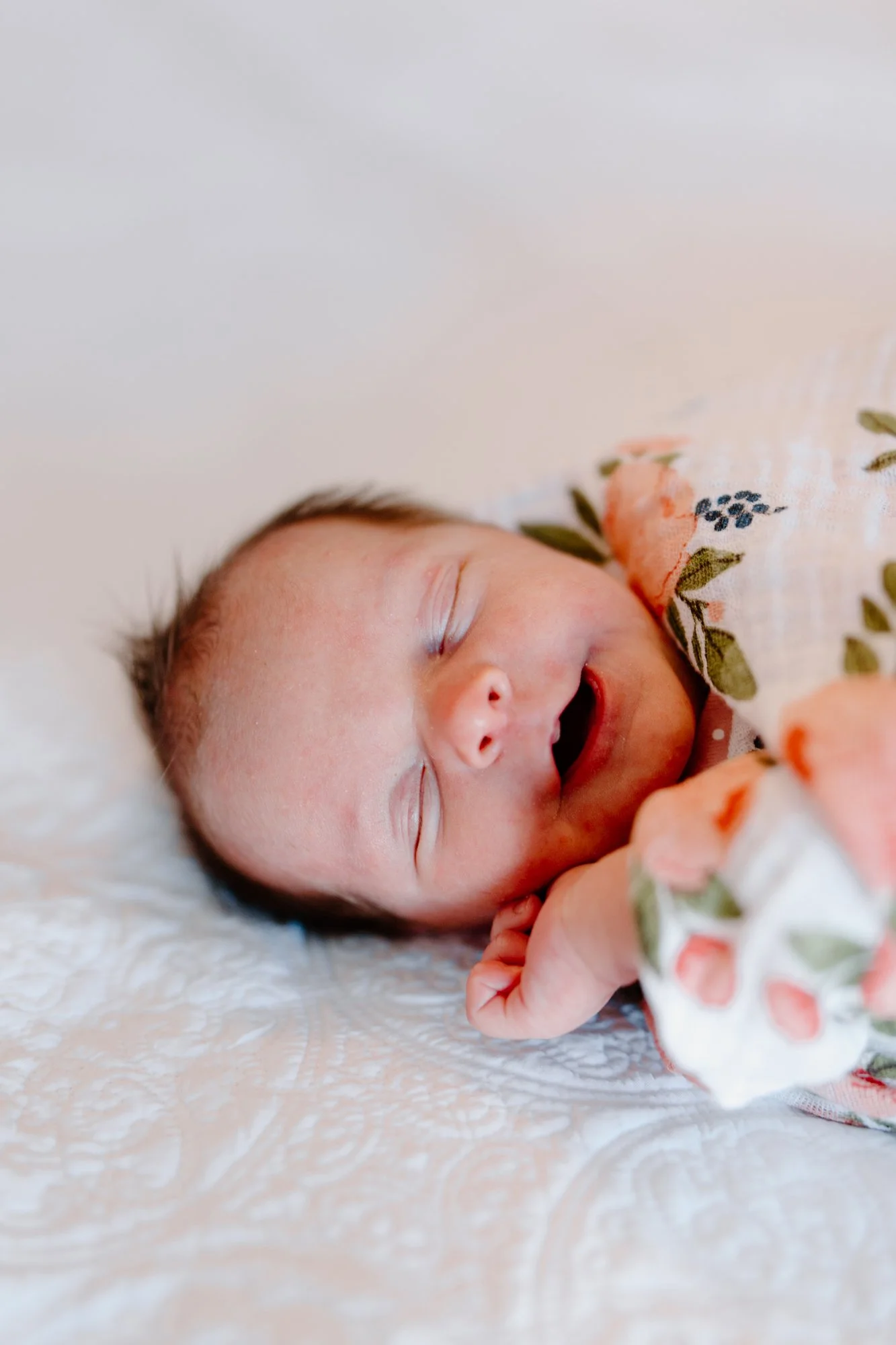 A smiling newborn baby sleeping on a white textured surface, dressed in a floral outfit.