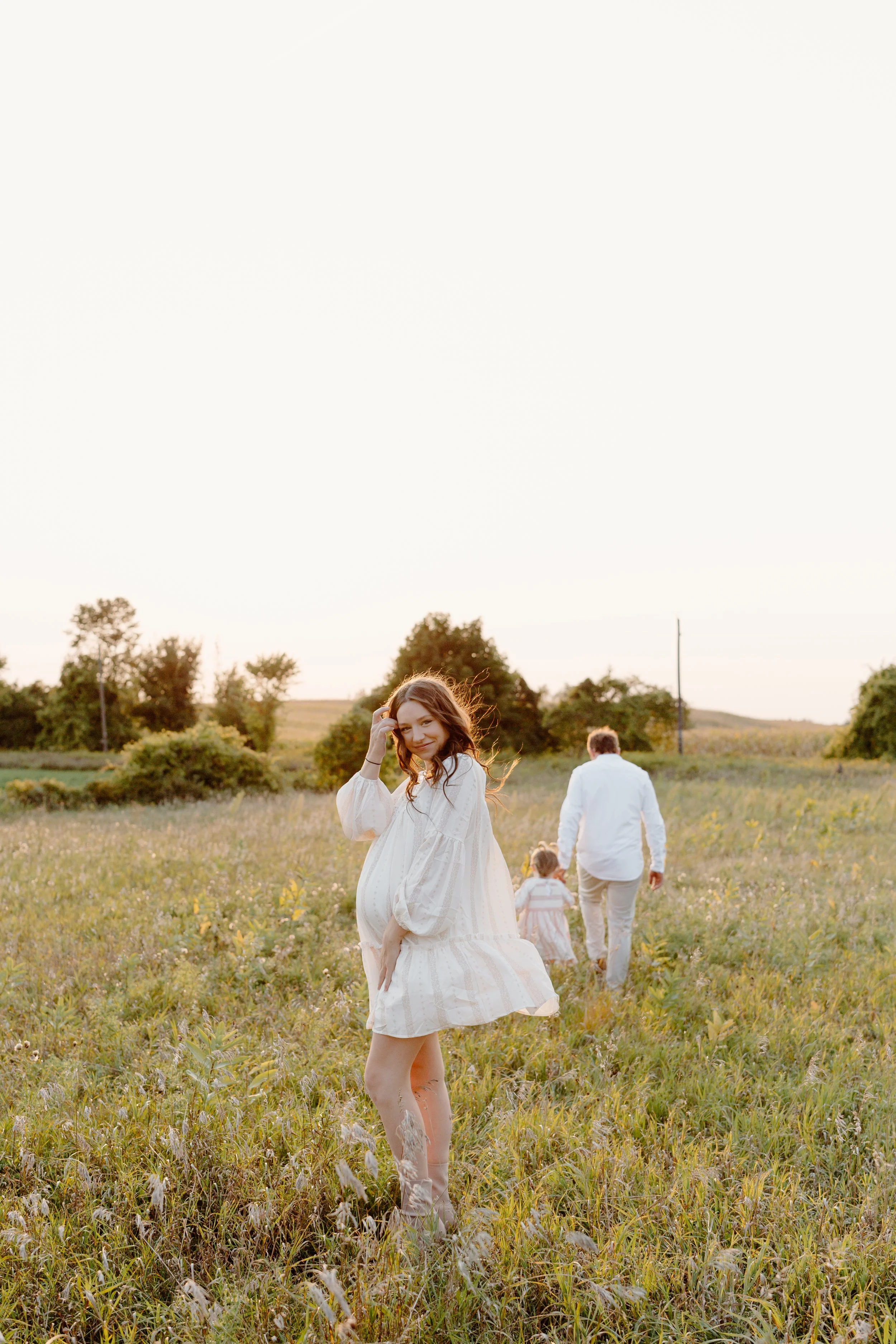 A woman in a white dress standing in a grassy field at sunset, with a man and two children walking away in the background.