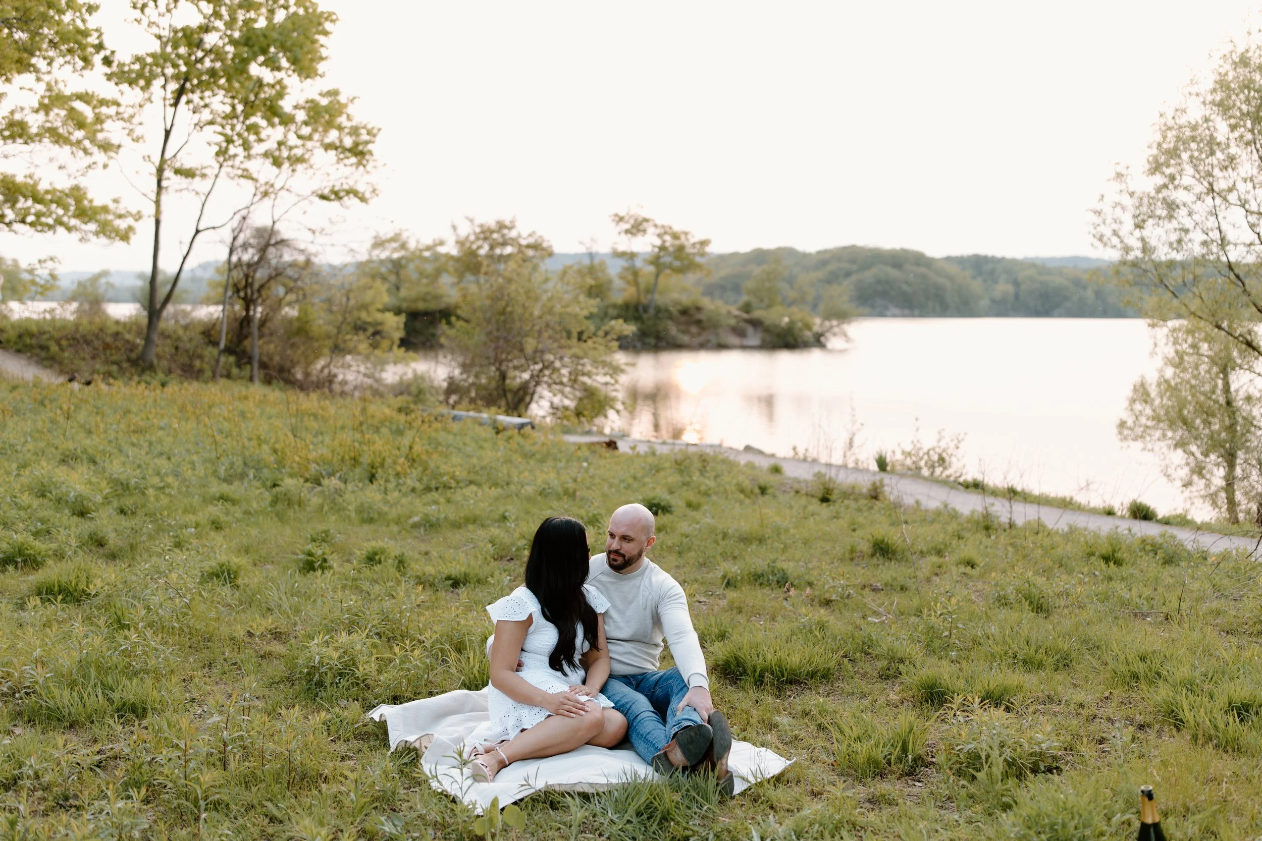 A couple sitting on a blanket near a river, surrounded by greenery and trees, with a bottle of champagne nearby.