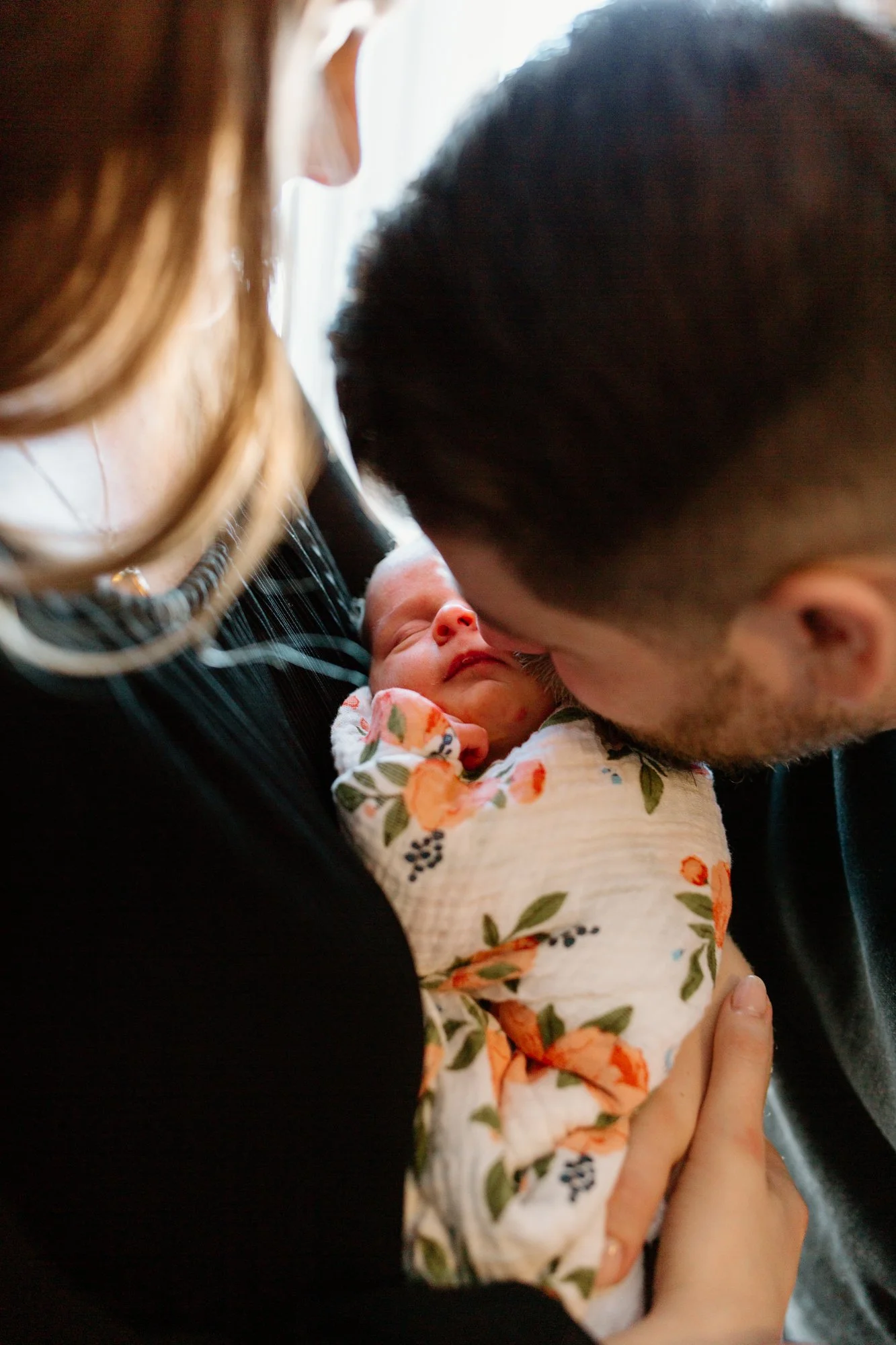 A woman and a man gently kiss a newborn baby wrapped in a floral blanket, holding and cradling the baby close.