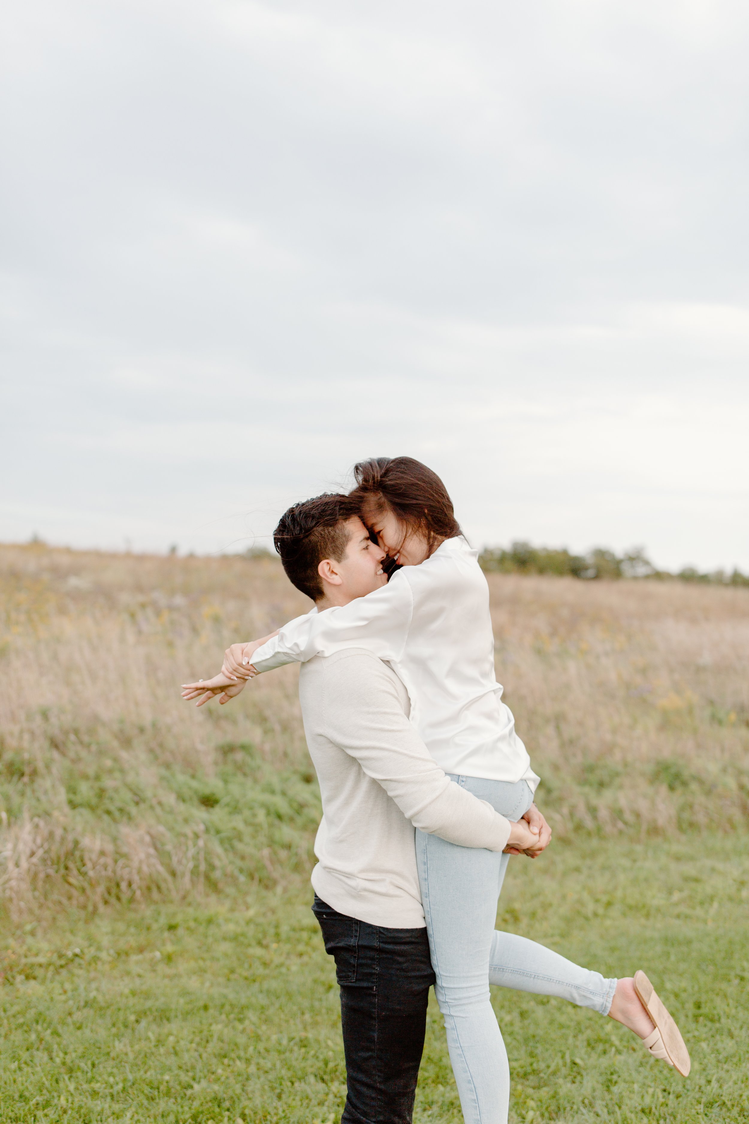 A couple embraces in a grassy field during daytime, with the woman lifting her legs off the ground and their foreheads touching affectionately.