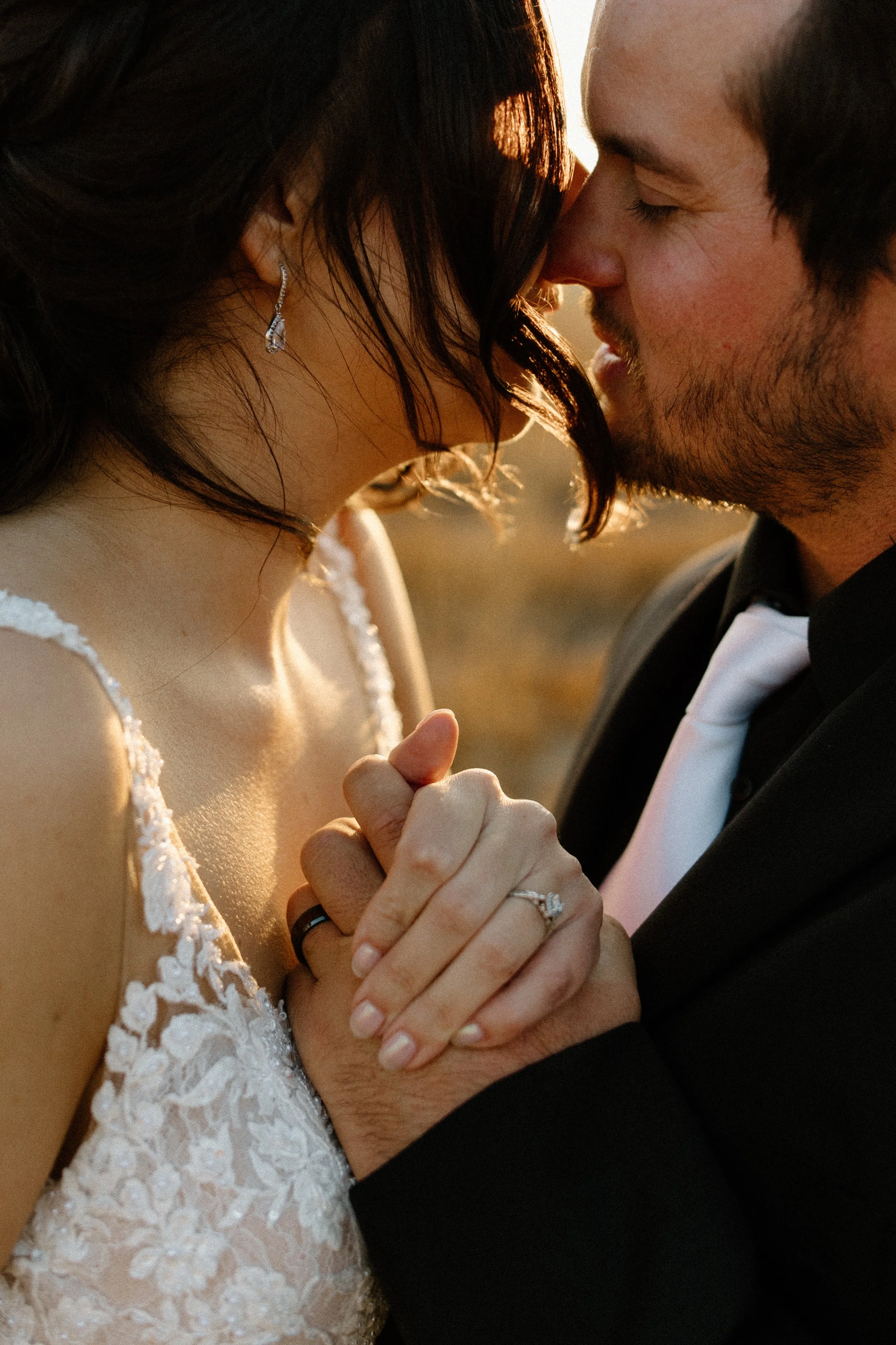 A couple dressed in wedding attire sharing a tender moment, with their faces close together and holding hands, on a warm, outdoor setting at sunset.
