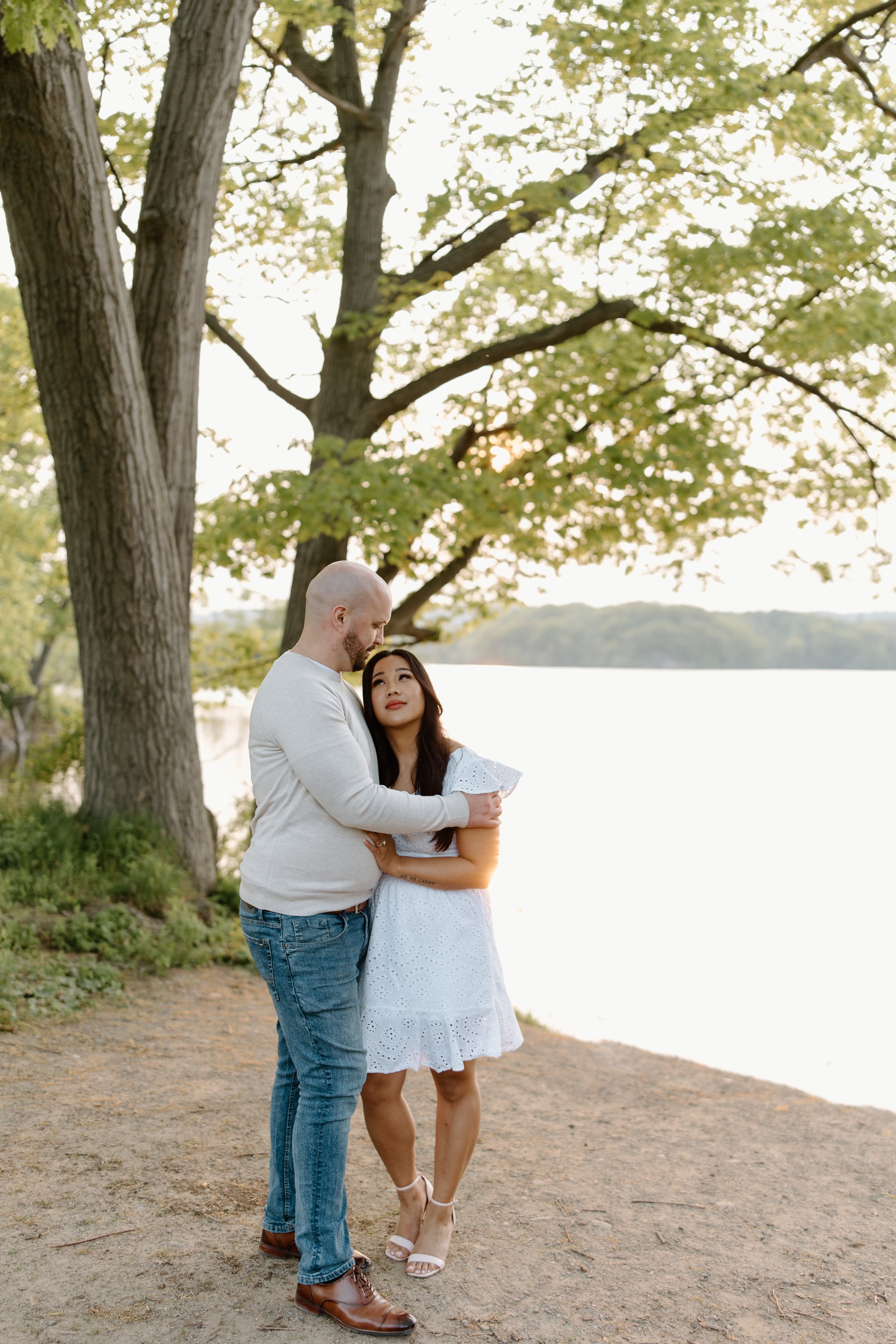 A couple embracing by a lake, with large trees and green foliage in the background, during sunset or late afternoon.