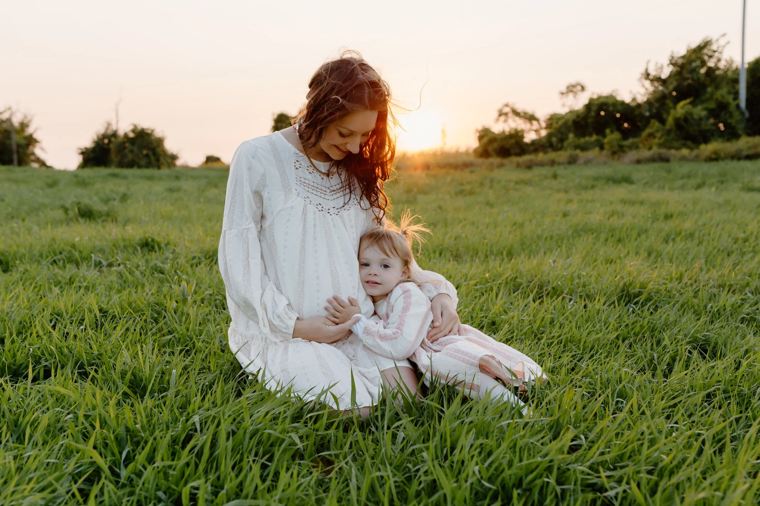 A pregnant woman and a young girl sitting together in a grassy field during sunset, with the woman smiling down at the girl and the girl hugging her belly.