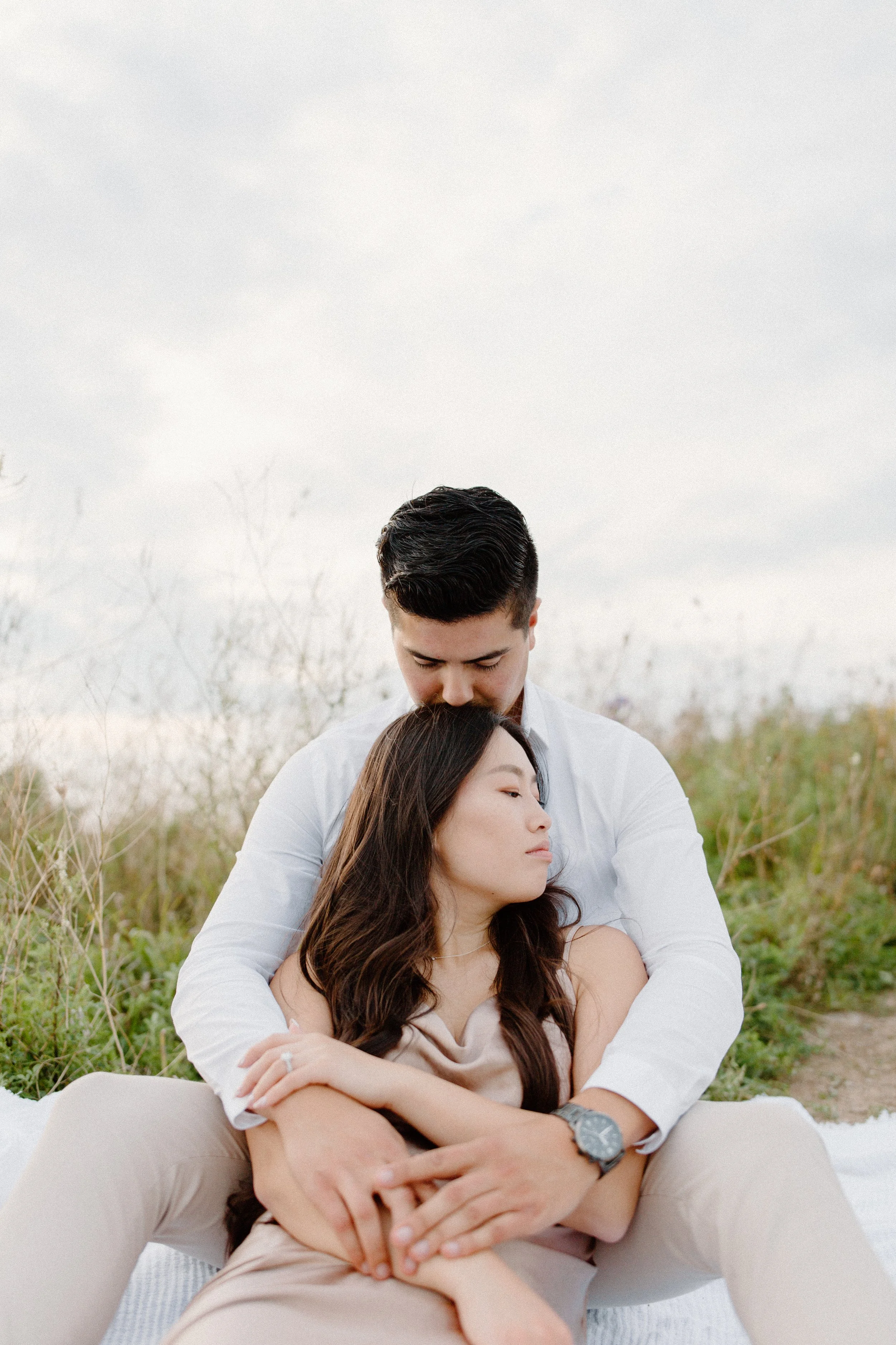 A couple sitting outdoors on a blanket, with the man hugging the woman close and resting his head against hers, in a natural setting with tall grass and an overcast sky.