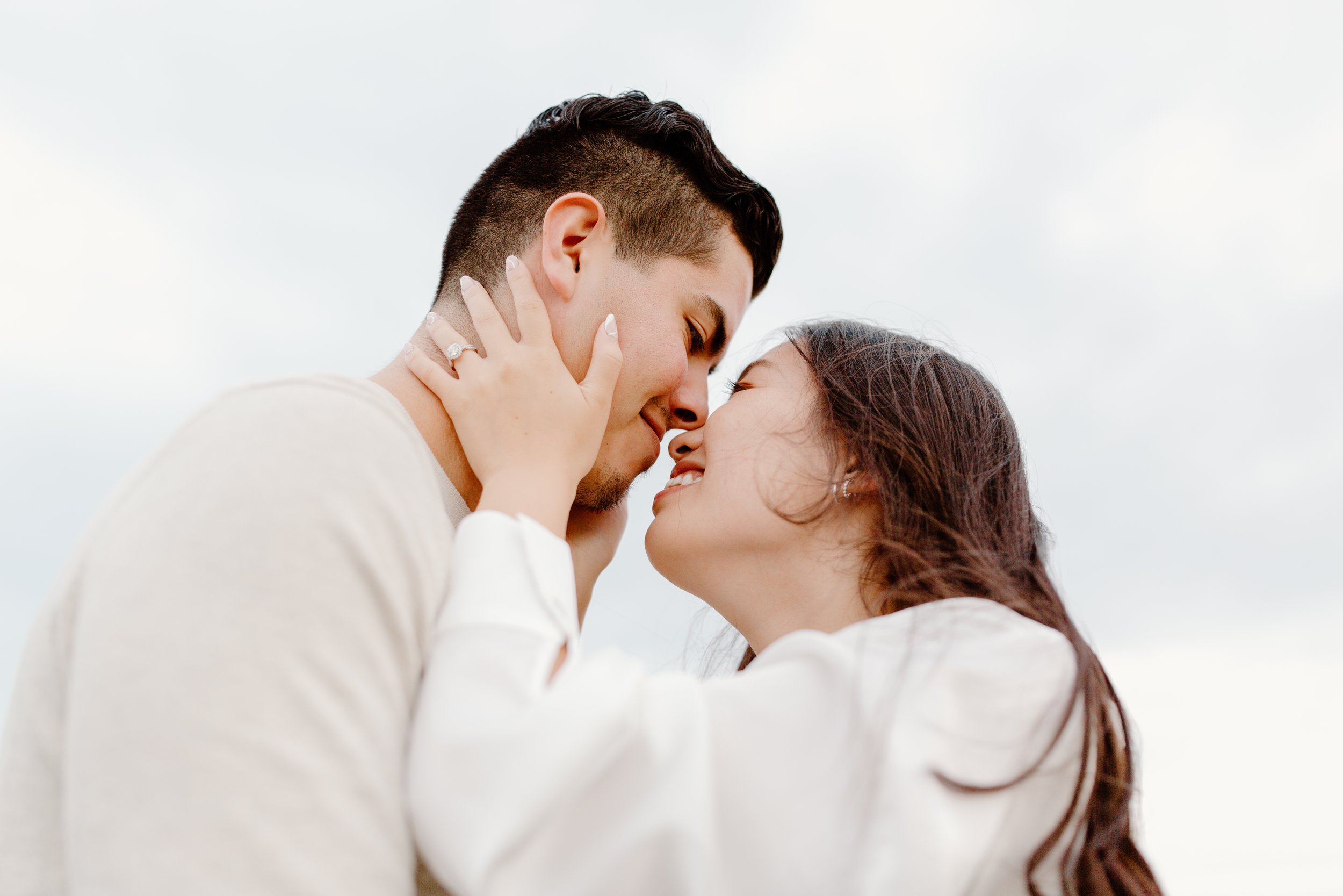 A couple standing close, touching foreheads, smiling, with the woman touching the man's face, against a cloudy sky.