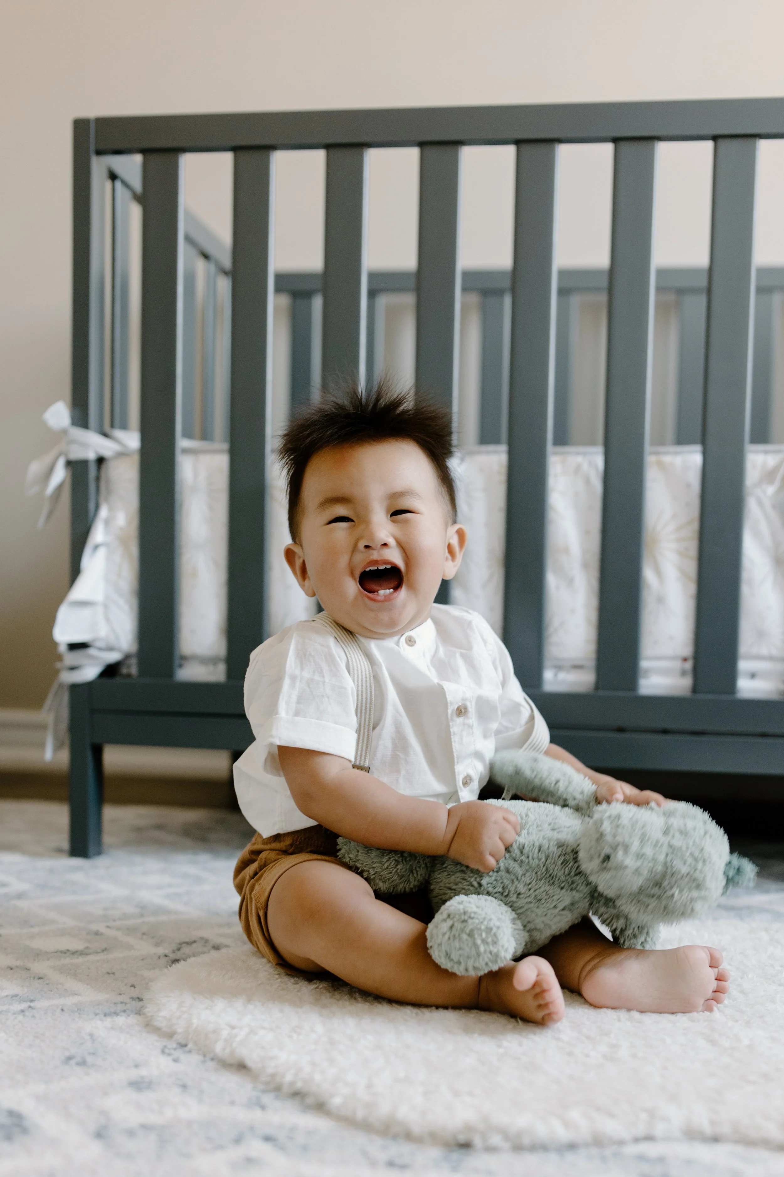 A laughing toddler sitting on a soft rug in front of a baby crib, holding a plush stuffed animal.