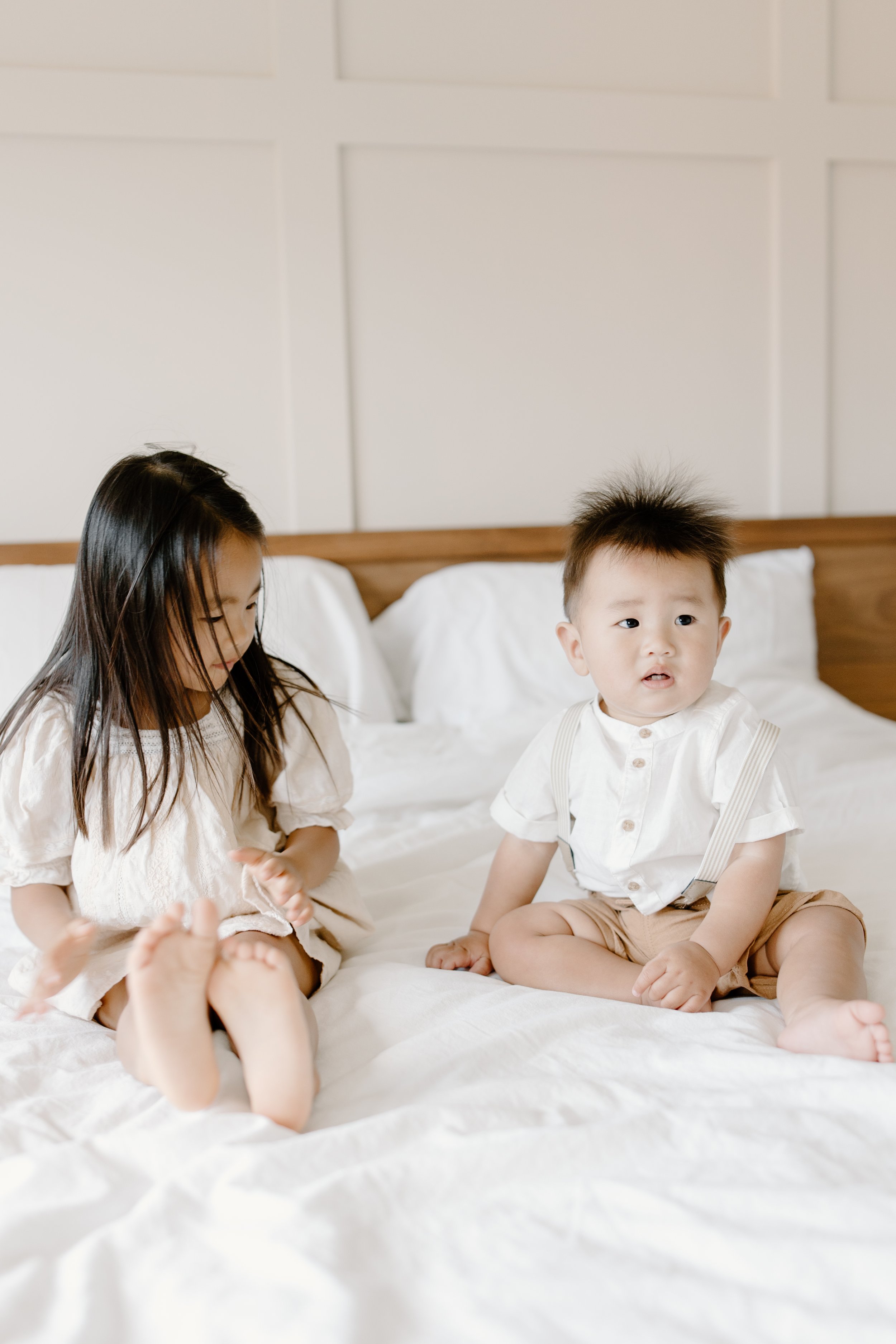 A young girl and a young boy sitting on a bed in a bright, modern room with white walls and bedding. The girl has long dark hair and is looking down, while the boy has short hair and is looking to the side with a curious expression.