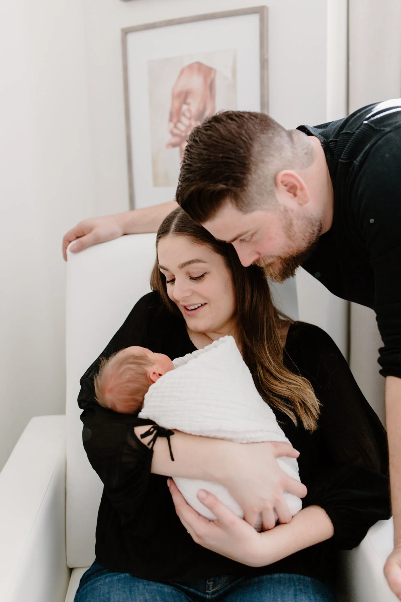 A woman sitting on a white chair holding a newborn wrapped in a white blanket, with a man leaning over and looking at the baby.