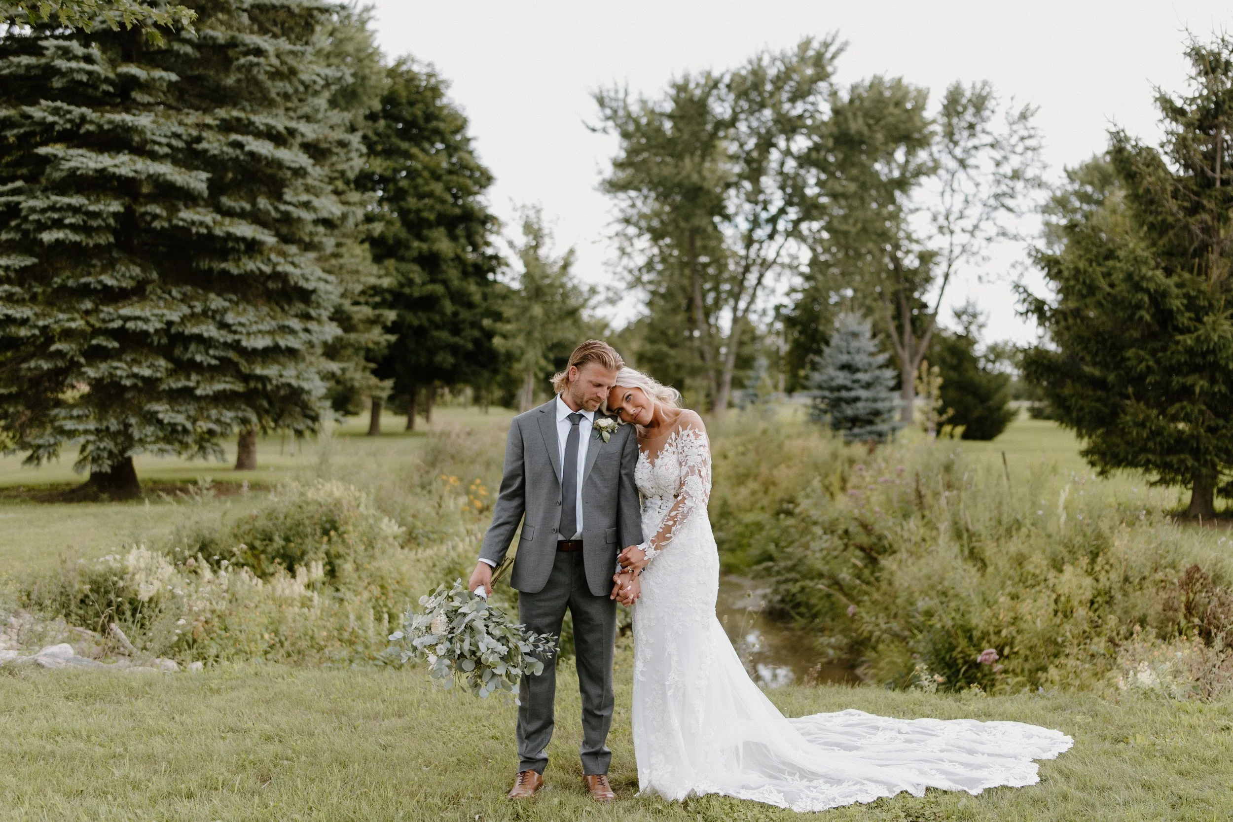 A bride and groom standing together outdoors on a grassy area near a small stream, surrounded by tall trees and greenery. The bride is wearing a white lace wedding dress and leaning her head on the groom's shoulder. The groom is dressed in a gray sui