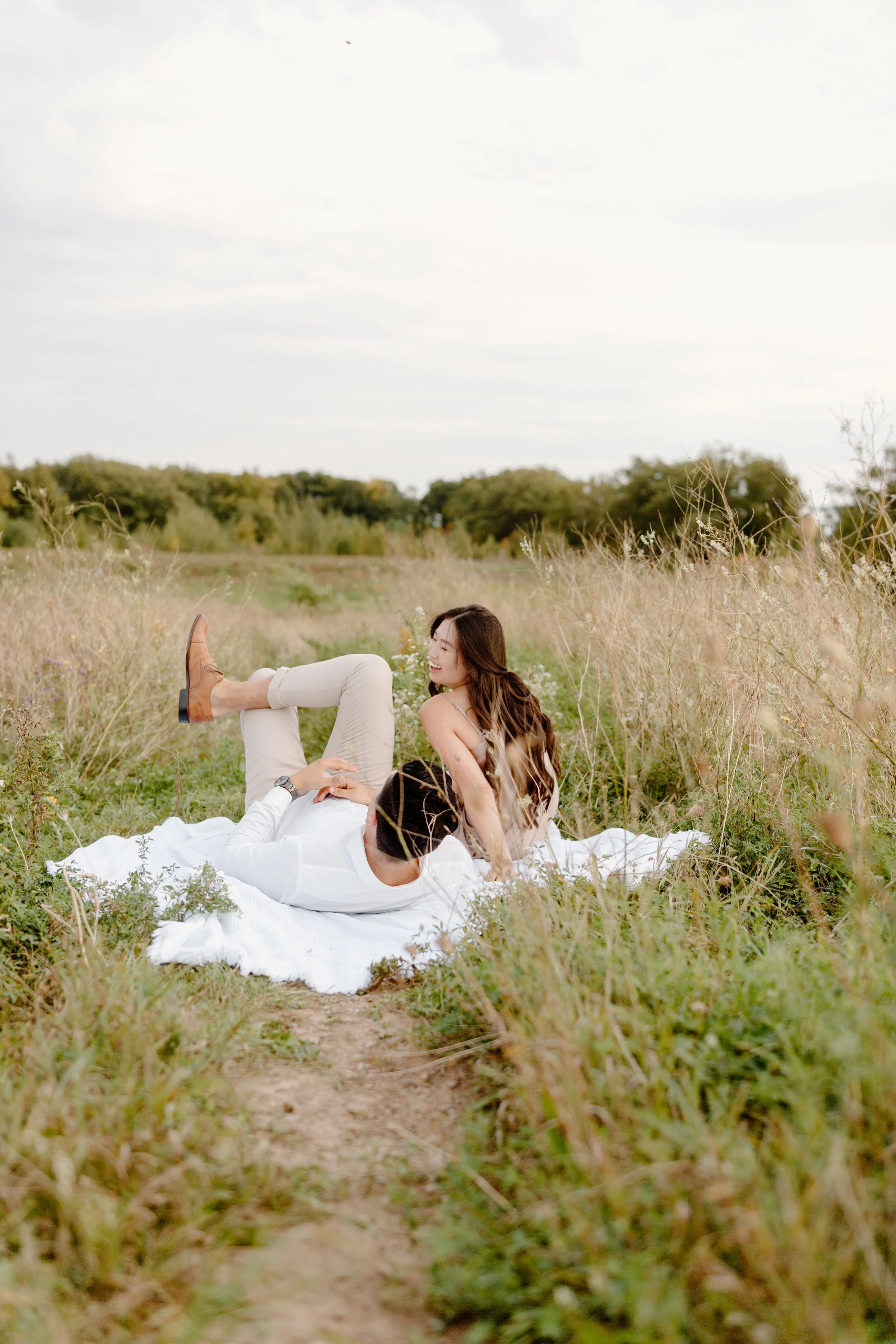 A couple relaxing on a white blanket in a grassy field, with the woman sitting and the man lying down, smiling and enjoying each other's company.