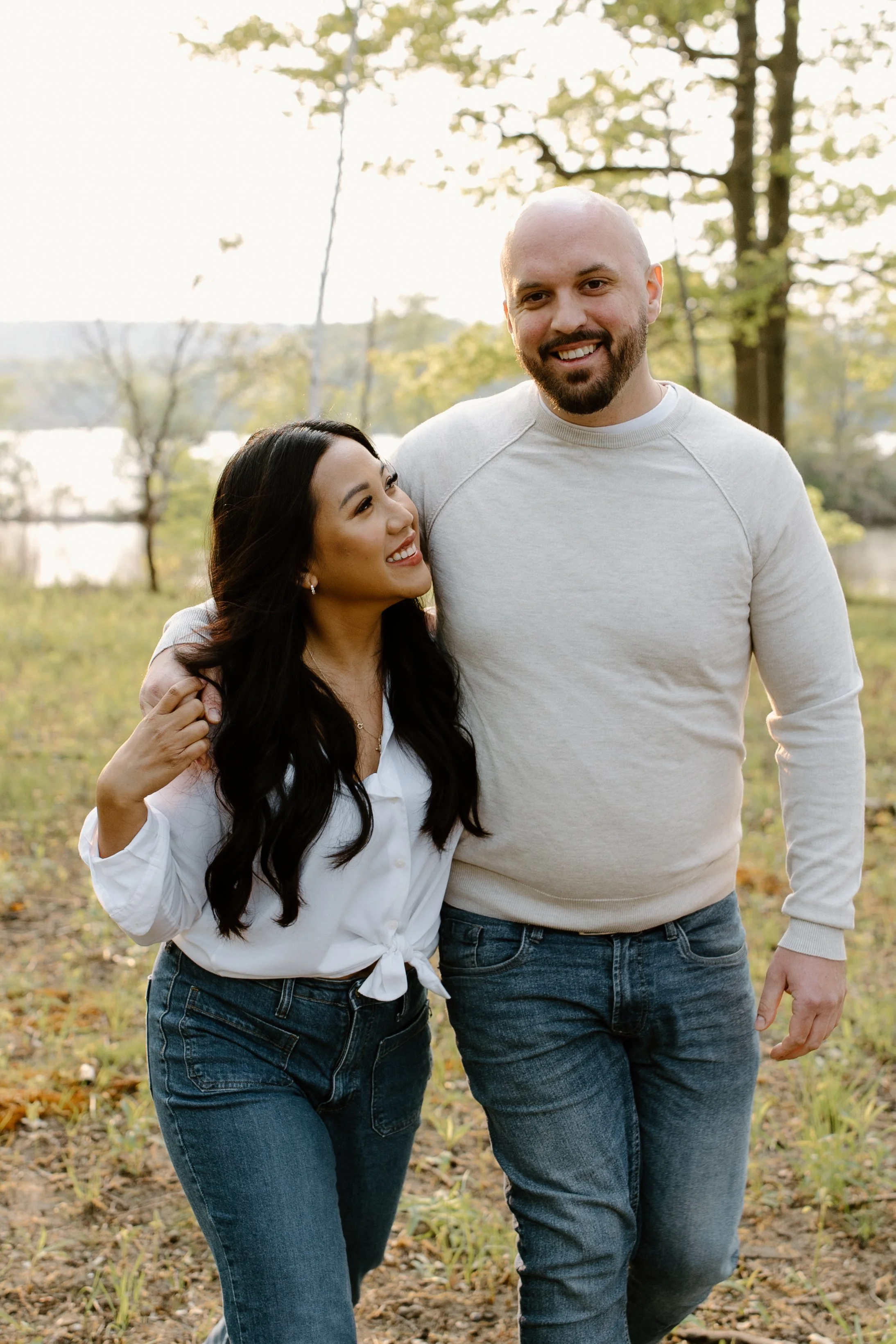 A smiling woman and man walking through a park during autumn, with trees and a lake in the background.
