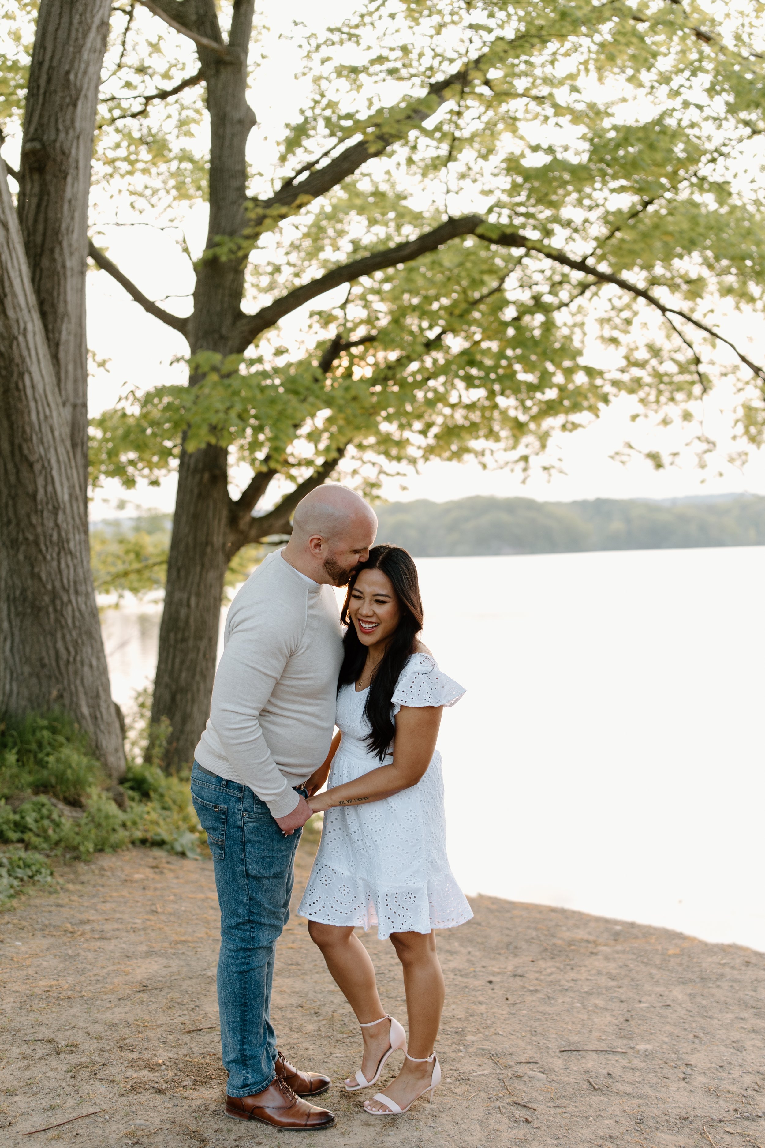 A happy couple standing close together outdoors near a lake, smiling and laughing under a large tree with green leaves, during daytime.