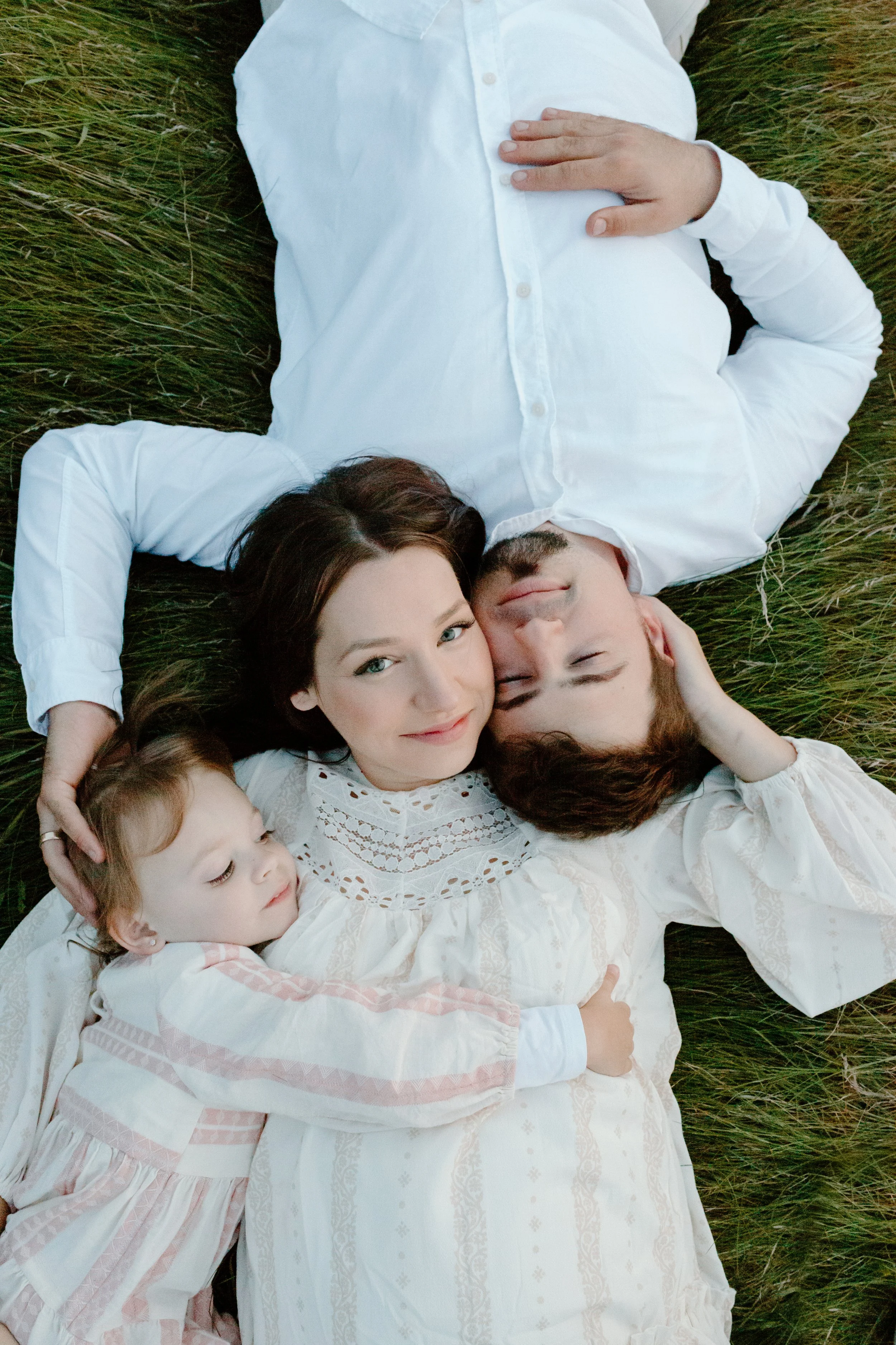 Family of three lying on grass, a mother, father, and young daughter, all dressed in white, cuddling and smiling.