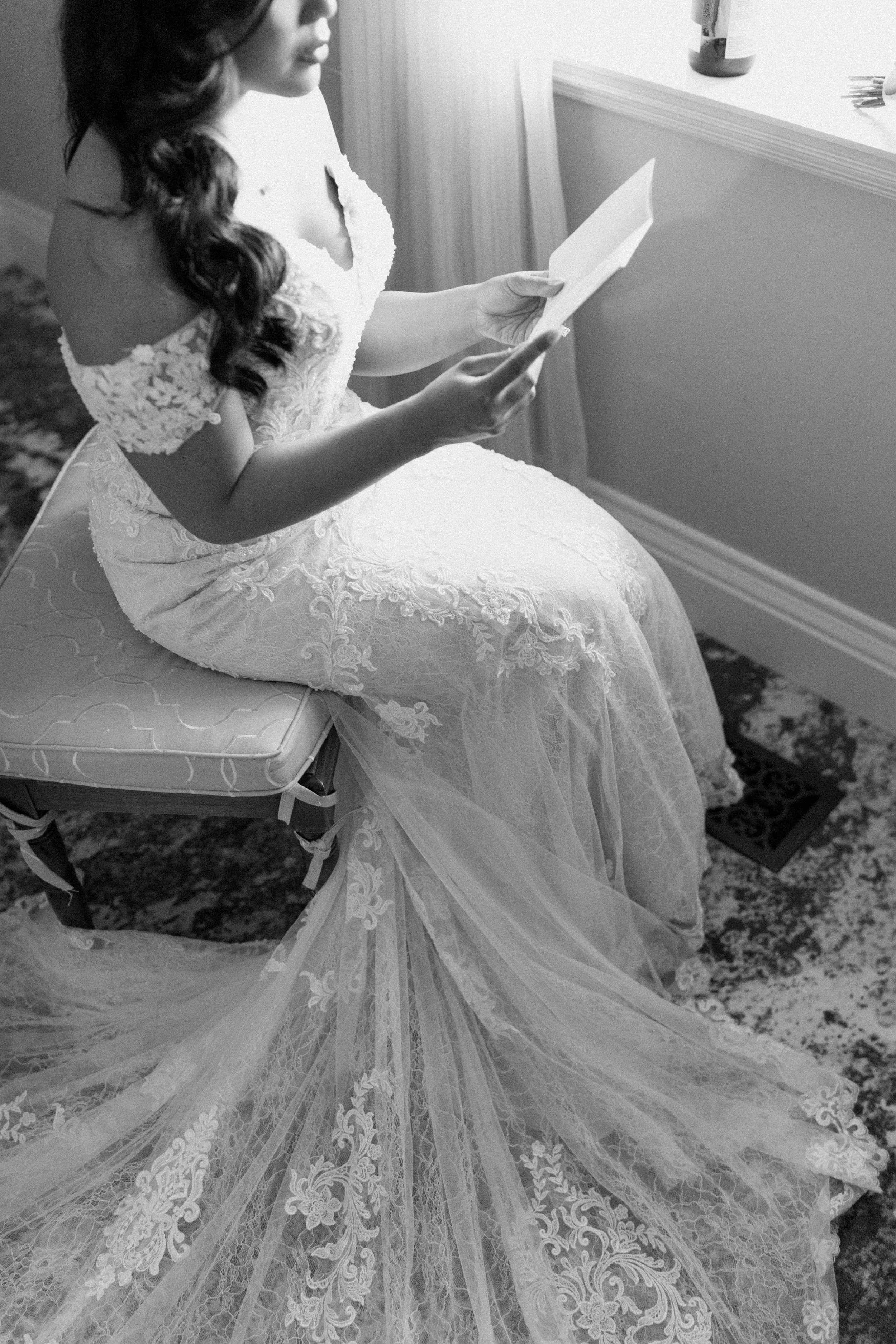 A woman in a wedding dress sitting on a chair, reading a card or note in front of a window.