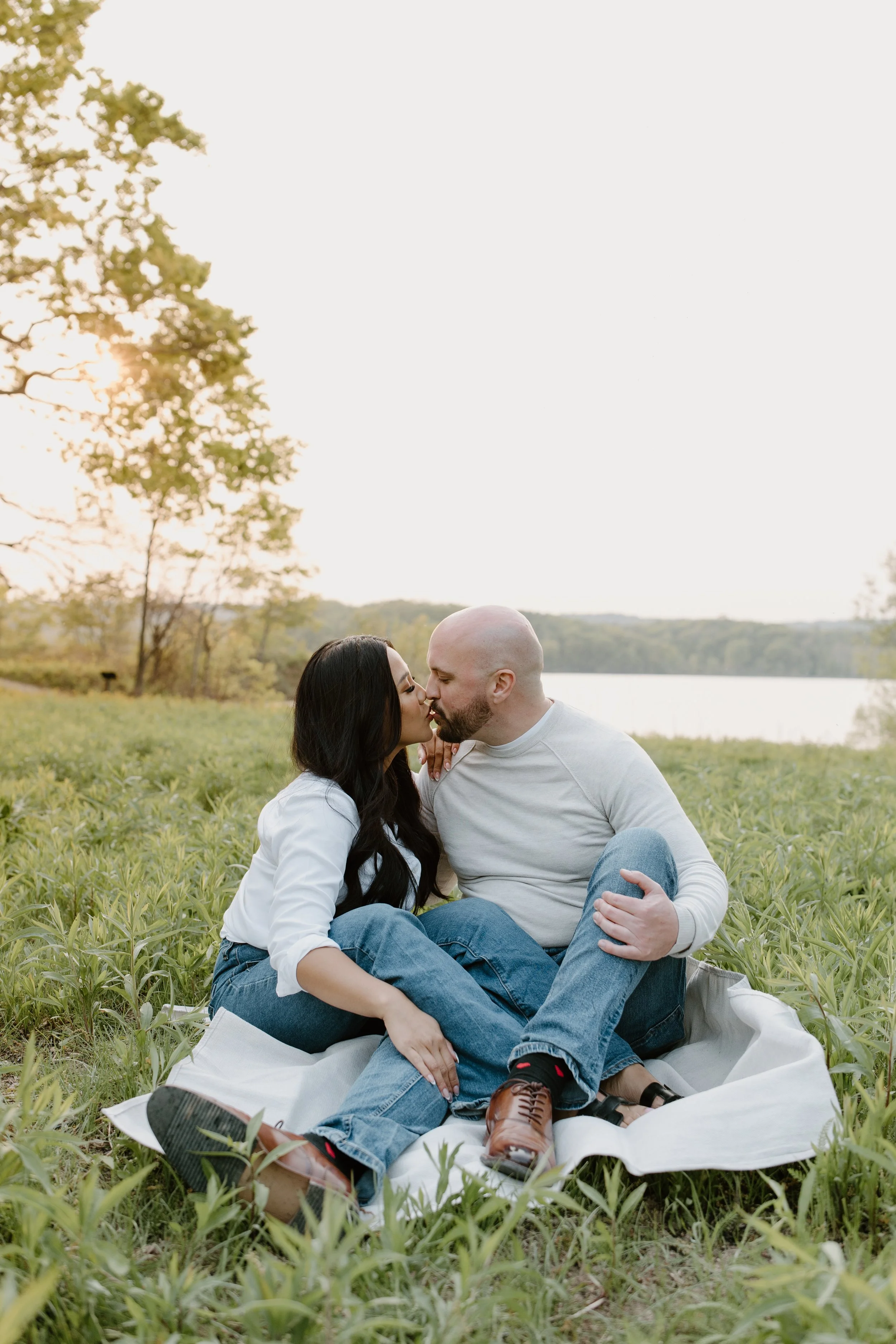 A couple sharing a kiss while sitting on a blanket in a grassy outdoor setting near a lake at sunset.