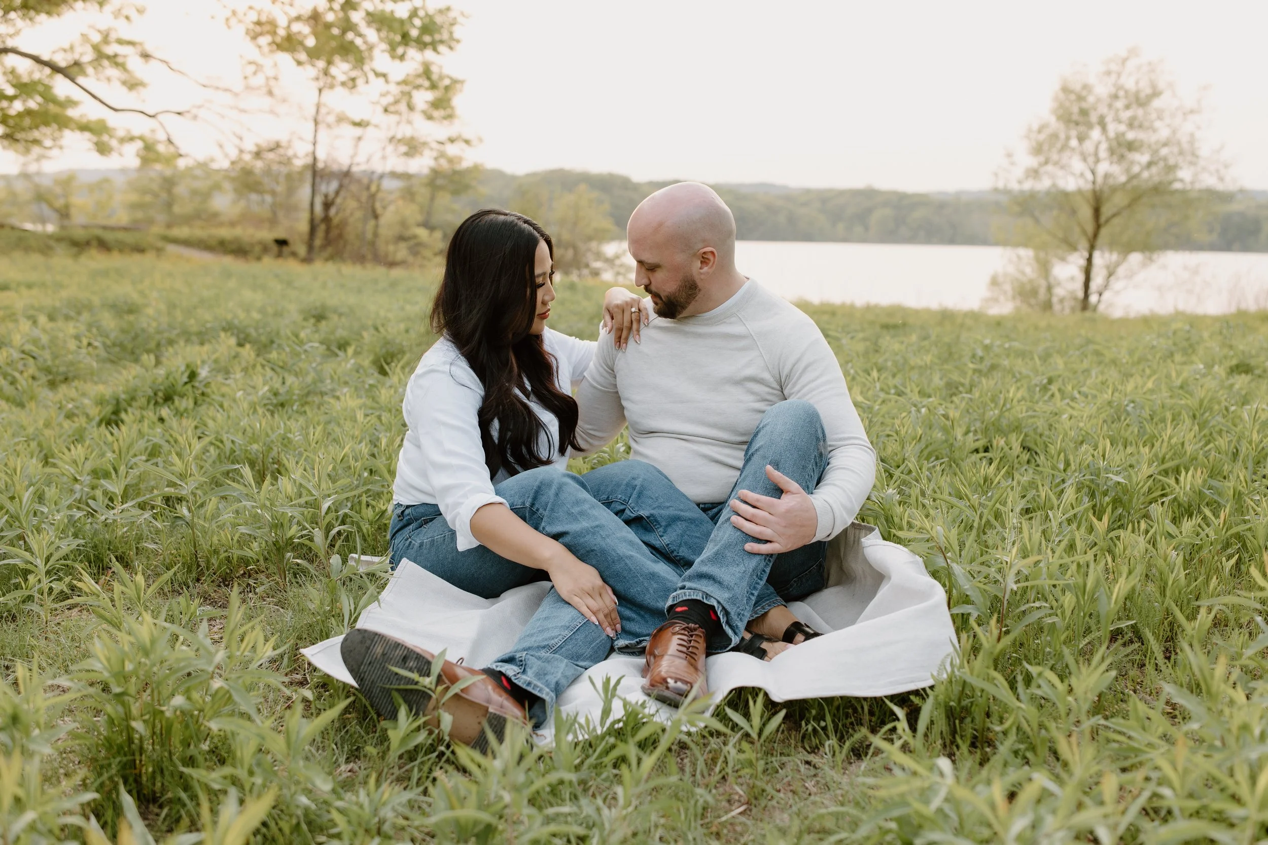 A couple sitting on a blanket in a grassy field near a lake, embracing and looking at each other.