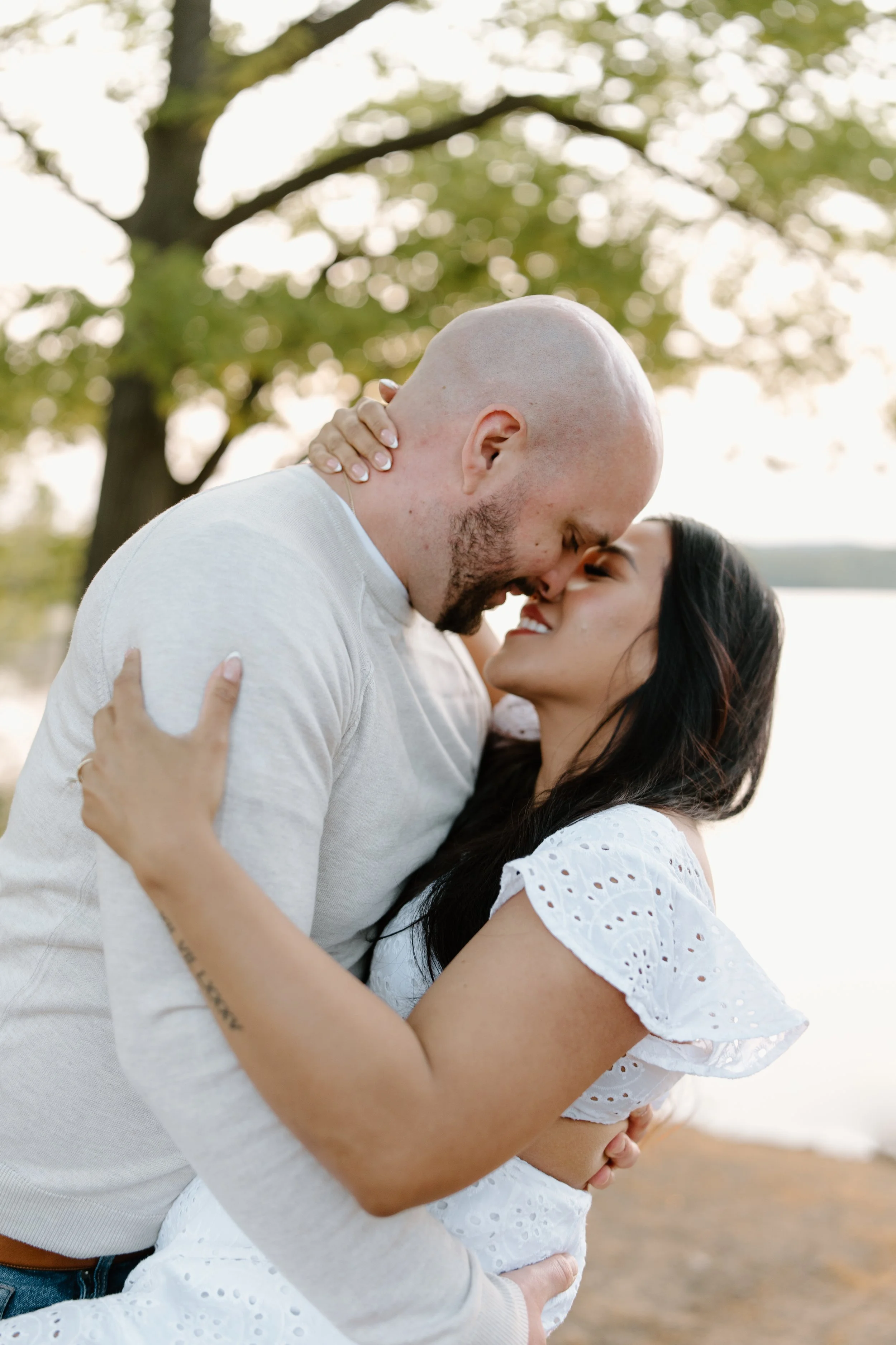 A couple embraces closely outdoors, with the man bald and the woman with dark hair, both smiling and about to kiss, near a body of water and trees.