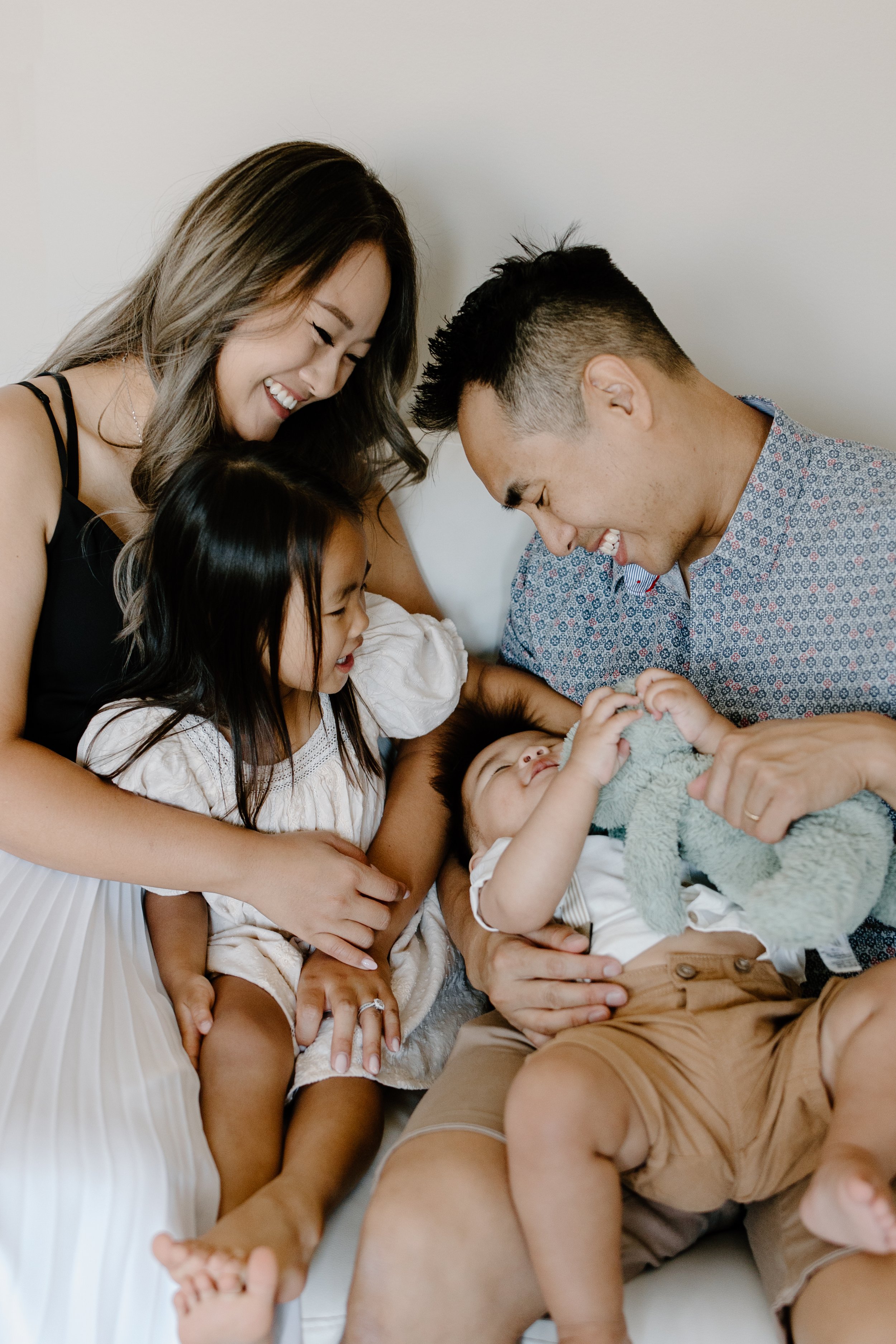 A multigenerational family of four smiling and playing together, with a woman, man, young girl, and baby on a white surface.