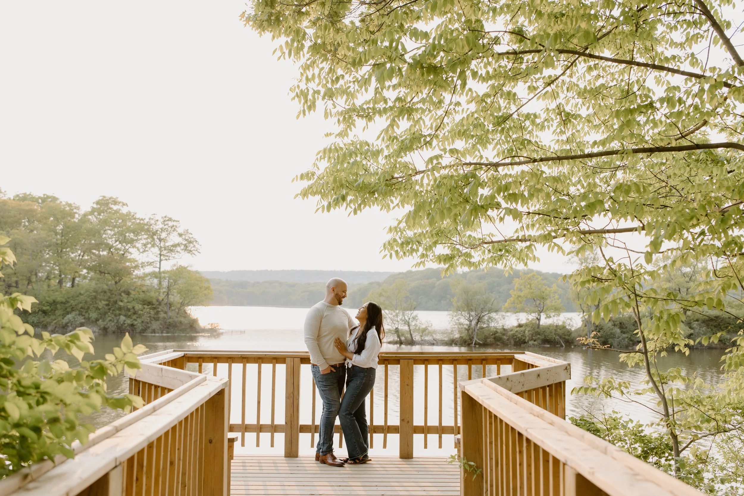 A couple standing on a wooden deck overlooking a lake, surrounded by trees, under a canopy of green leaves, during what appears to be late afternoon or early evening.