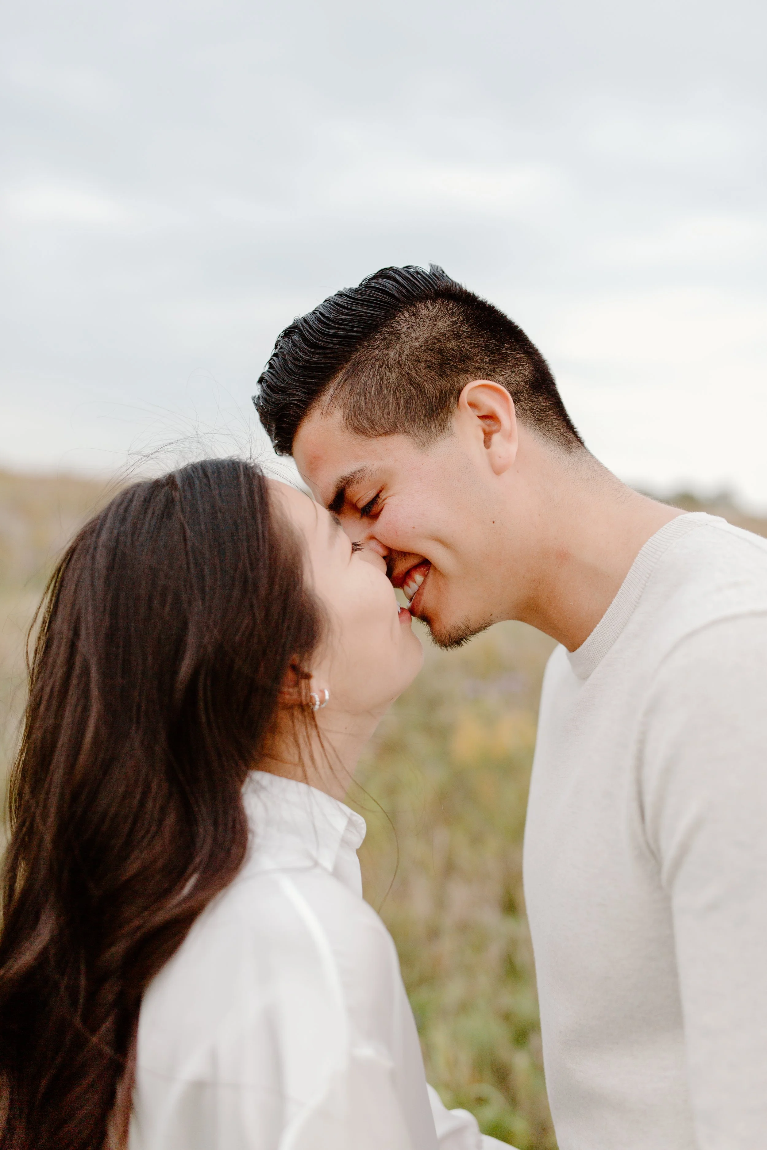 A young couple standing outdoors, touching foreheads and smiling, with a blurred natural landscape in the background.