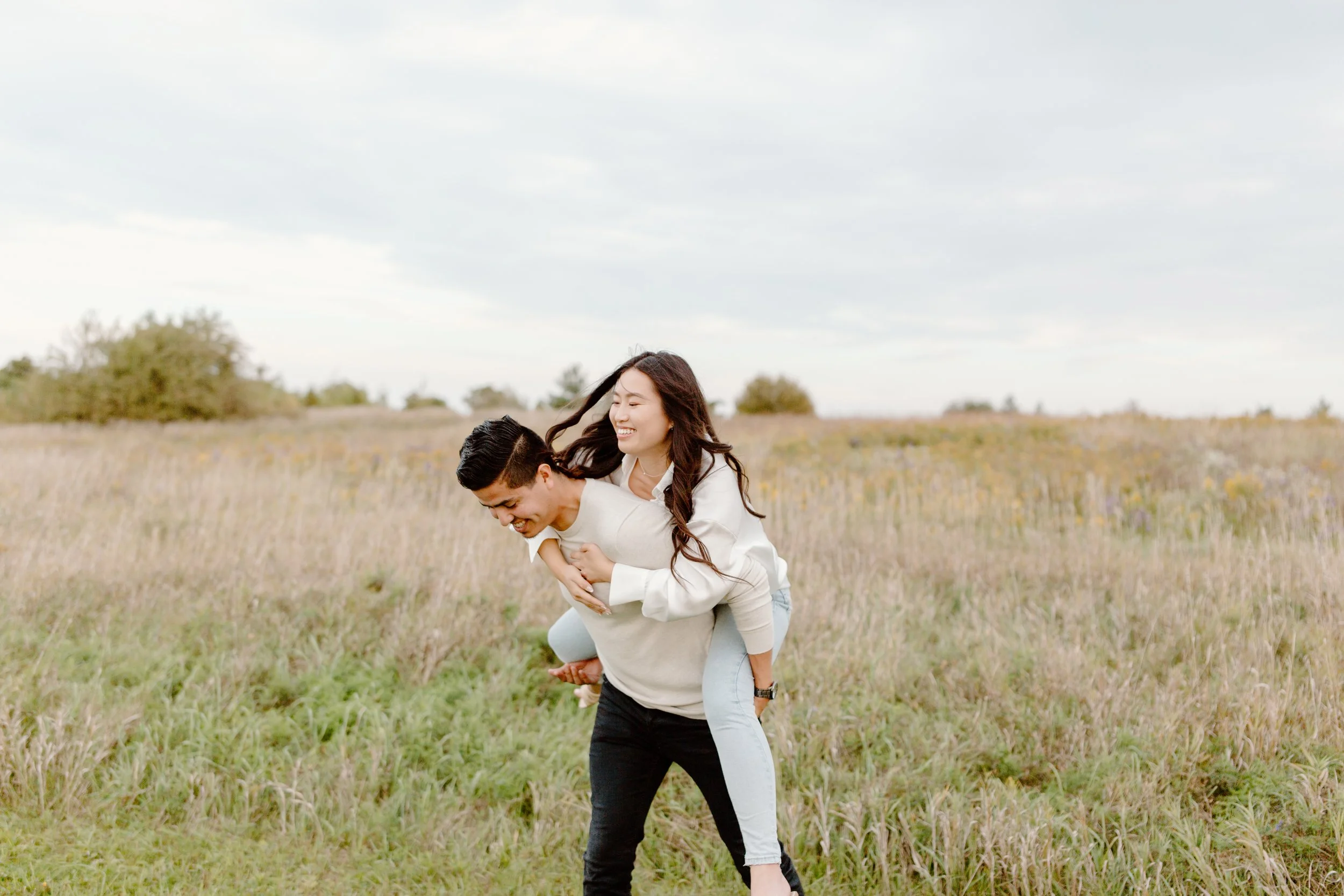 A young man giving a piggyback ride to a smiling young woman in an open grassy field with trees in the background, under a cloudy sky.