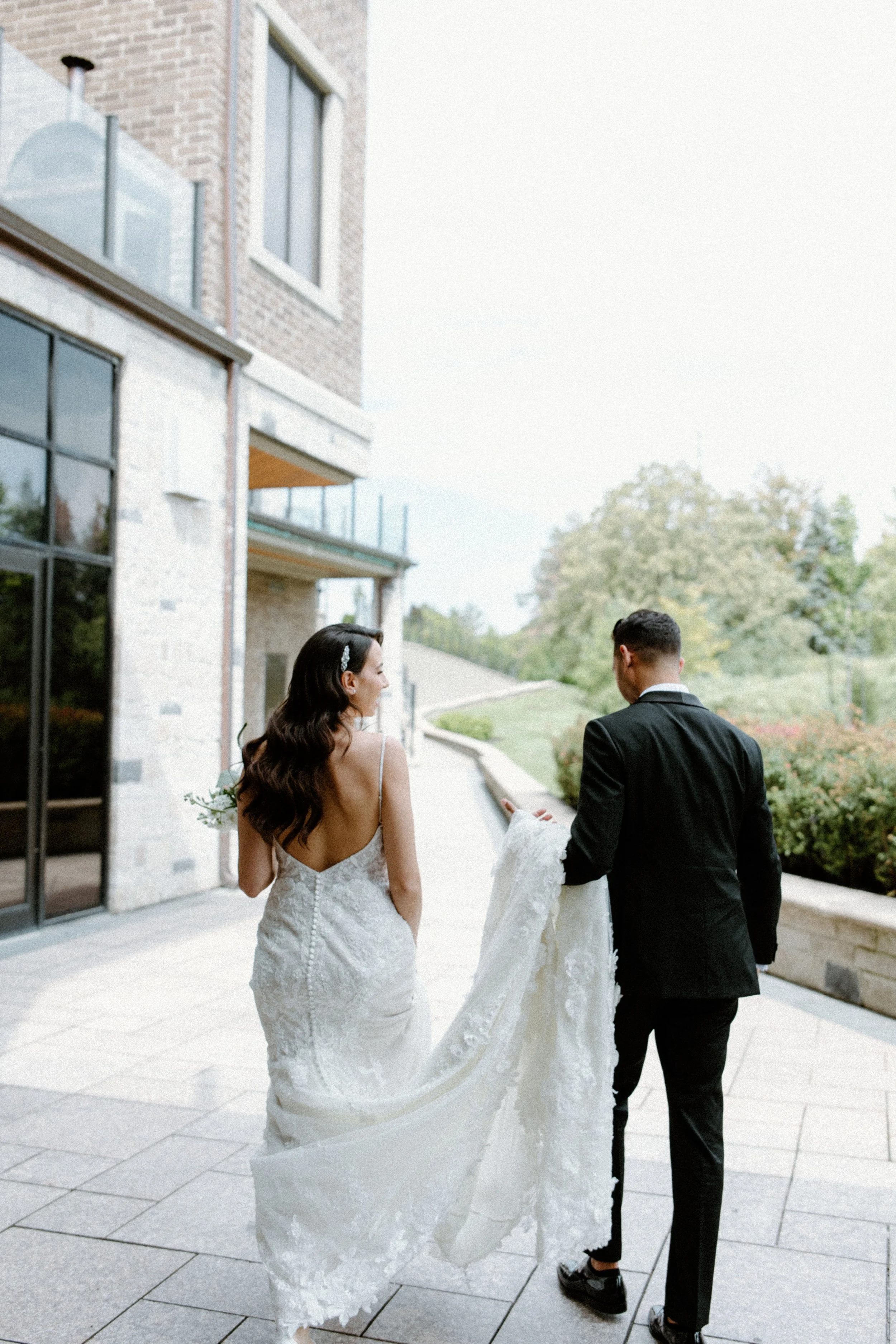 A bride and groom walk outdoors, the bride in a white wedding dress and the groom in a black suit, holding the bride's train, on a pathwayside with modern building and greenery around.